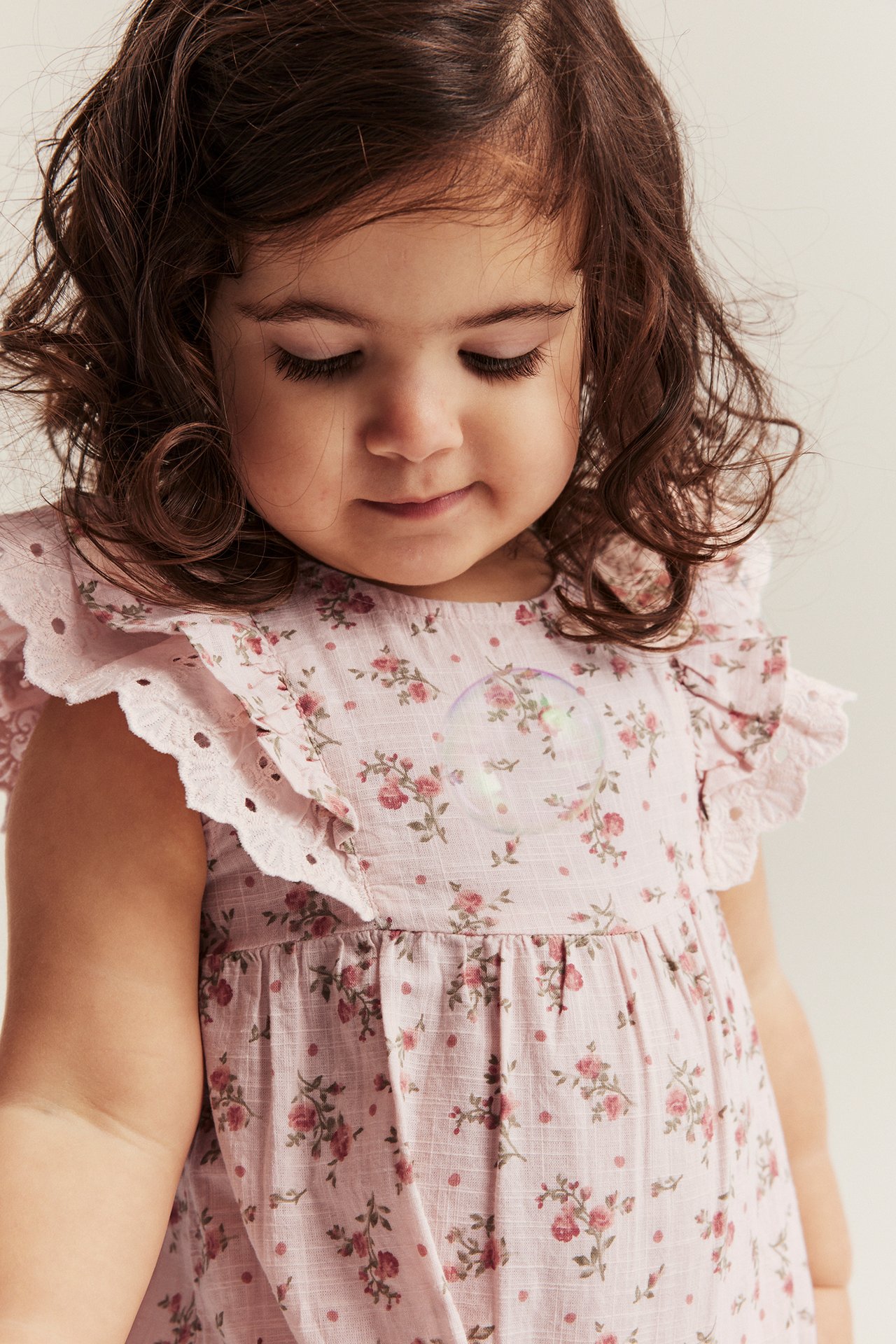Close-up front view: baby in a light pink floral cotton playsuit with ruffle lace sleeves, a bubble floats.