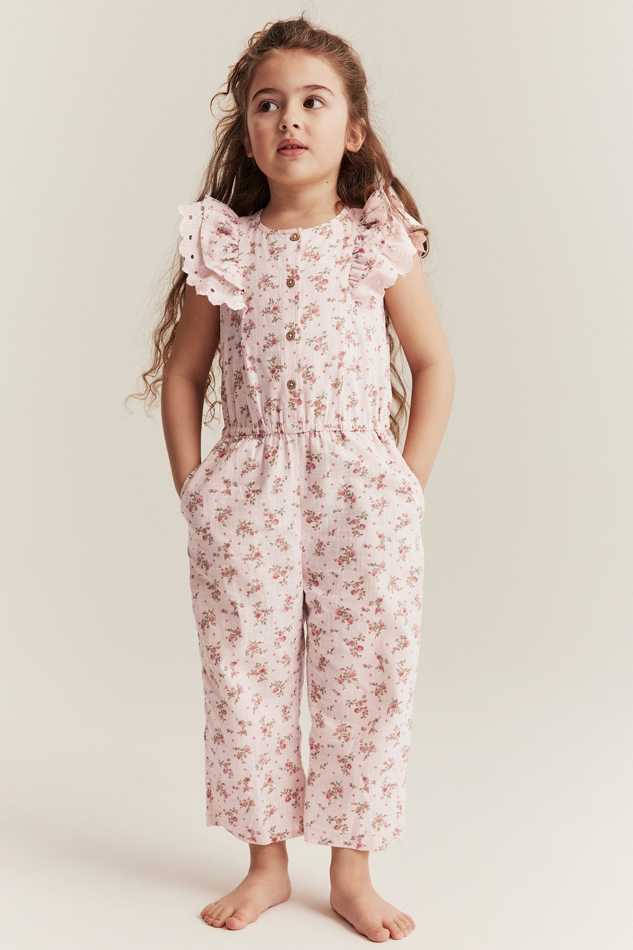 Vorderansicht eines Kindes in hellrosa geblümtem Baumwoll-Jumpsuit mit Rüschenärmeln, Knöpfen und Seitentaschen.