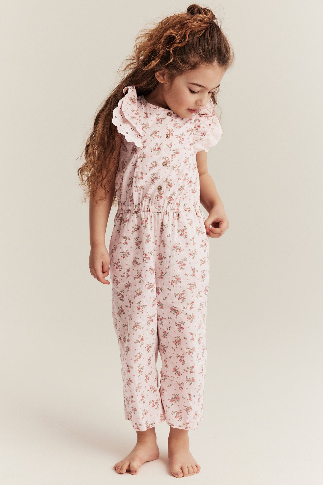 Vorderansicht: Kind in hellrosa geblümtem Jumpsuit mit Rüschenärmeln, Knöpfen vorne und Gummizug in der Taille.