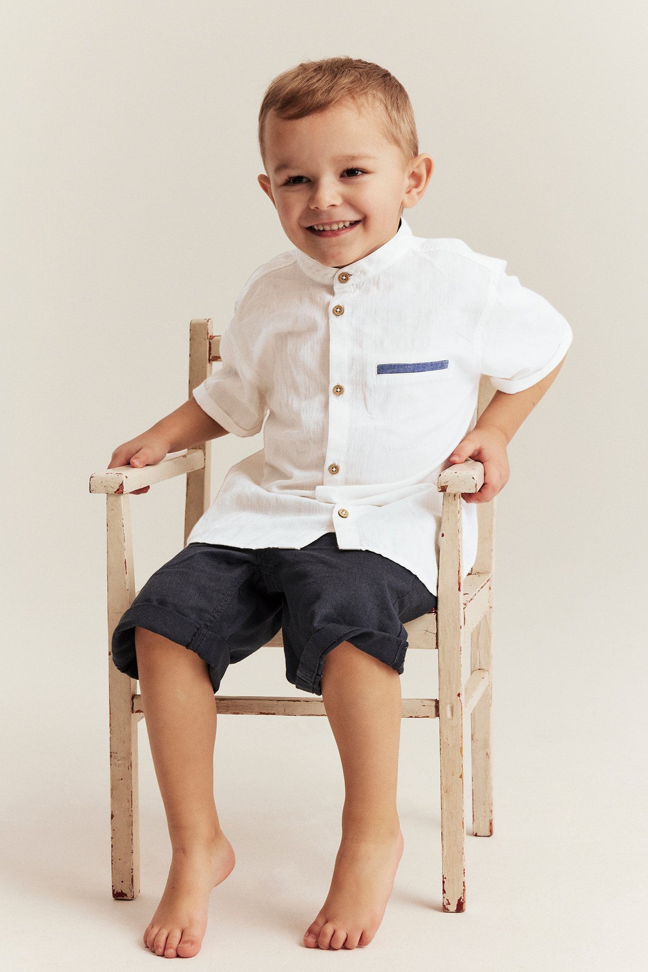 Vorderansicht eines Kindes in einem cremeweißen Webhemd mit kurzen Ärmeln, Kokosknöpfen, Chambray-Tasche und dunklen Shorts.