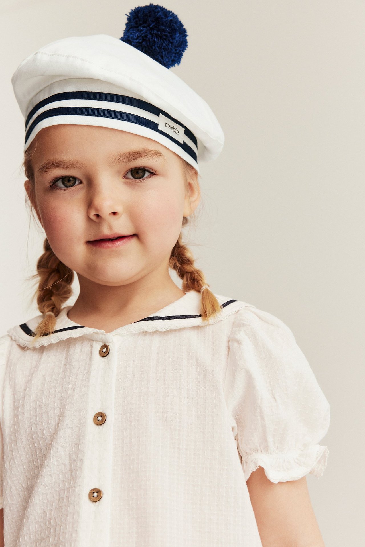 Vorderansicht eines Kindes in cremeweißer Baumwollbaskenmütze mit blauen Streifen und Bommel, über cremeweißer strukturierter Bluse.