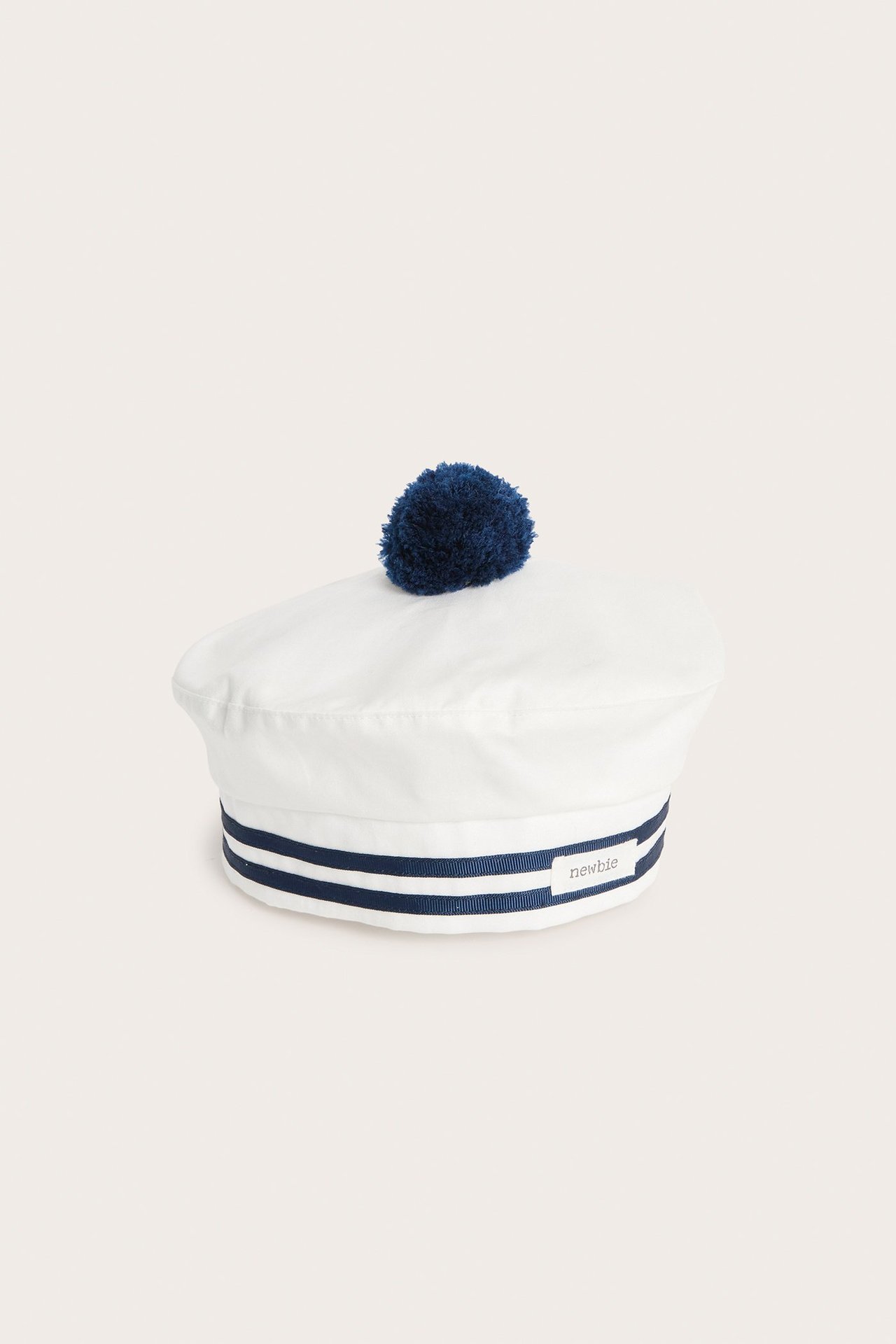 Vorderansicht einer cremeweißen Baby-/Kinder-Baskermütze aus Baumwolle mit dunkelblauen Streifen und Bommel.