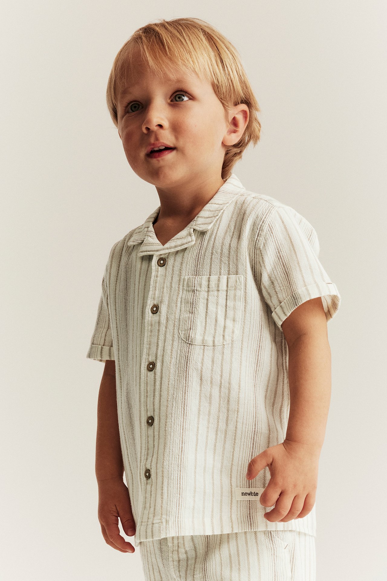 Vorderansicht: Kind in gestreiftem, kurzärmeligem Offwhite-Baumwollhemd mit braunen Knöpfen und passenden Shorts.