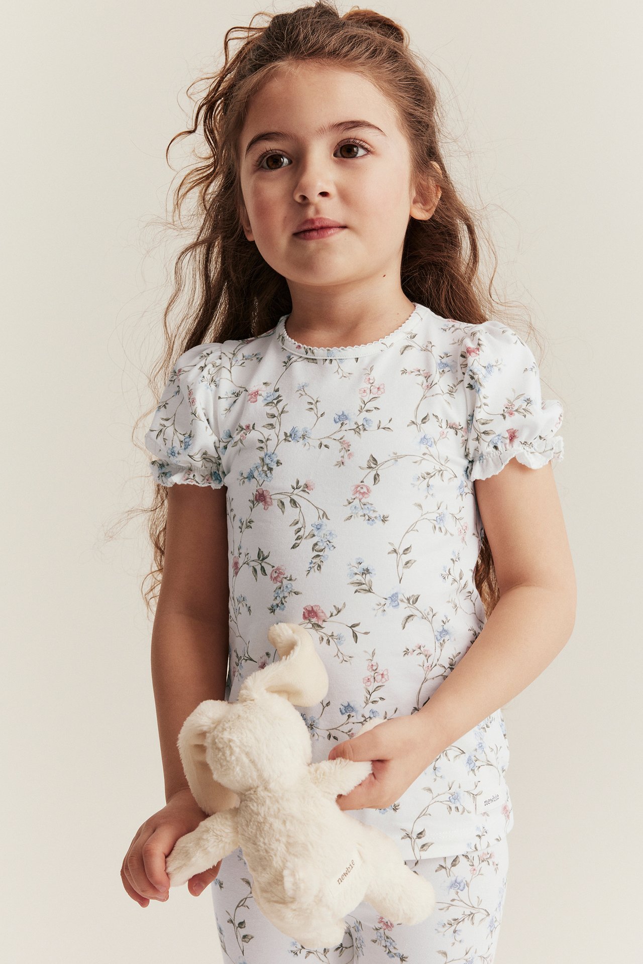 Front view: Kid in off-white floral short-sleeved top, matching trousers, holding a cream plush bunny.