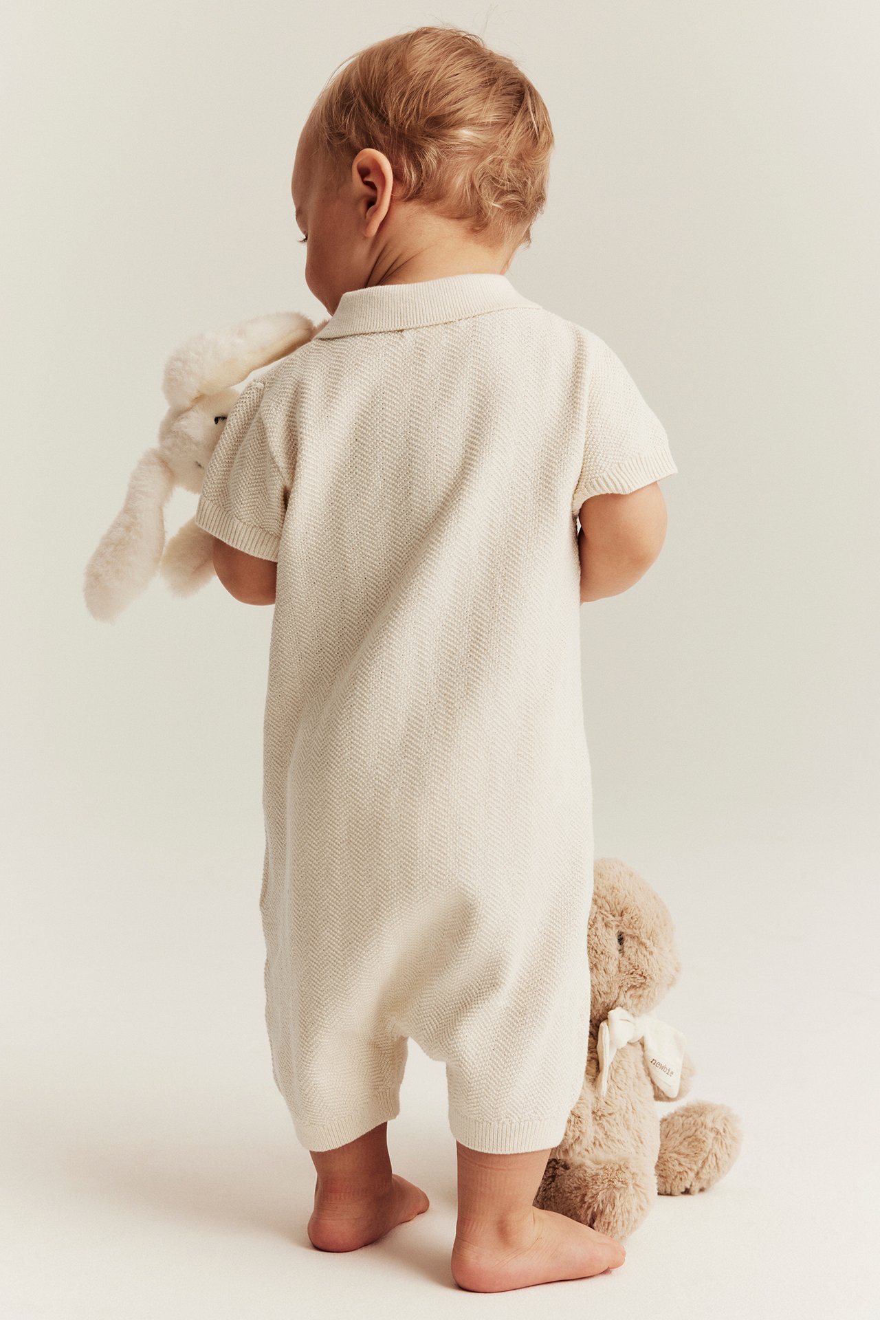 Back view: baby in a light beige knitted short-sleeved playsuit, holding a white bunny. Brown bear nearby.