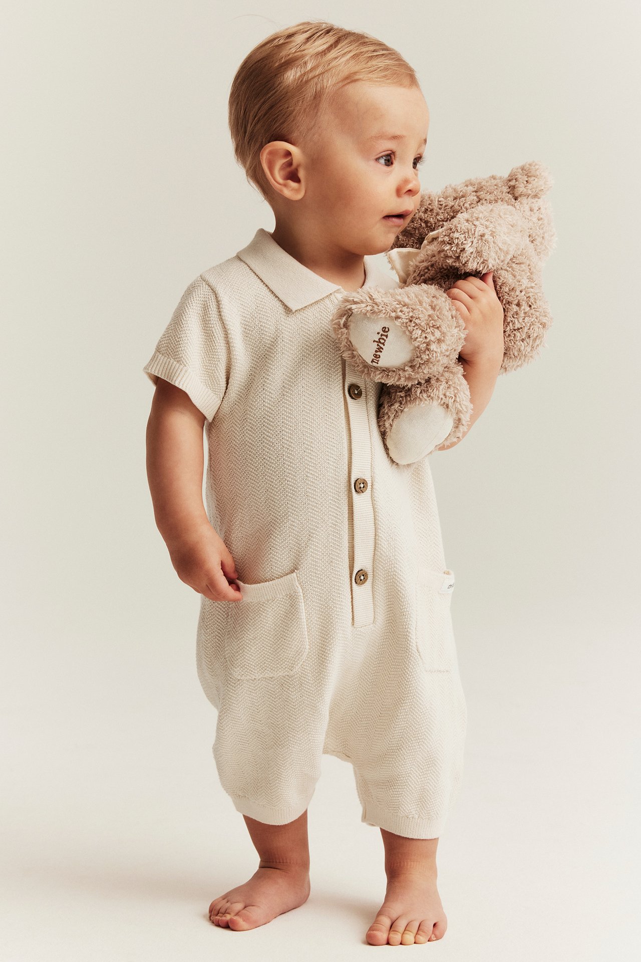 Side view of a baby in a light beige knitted short-sleeved playsuit, holding a brown teddy bear.
