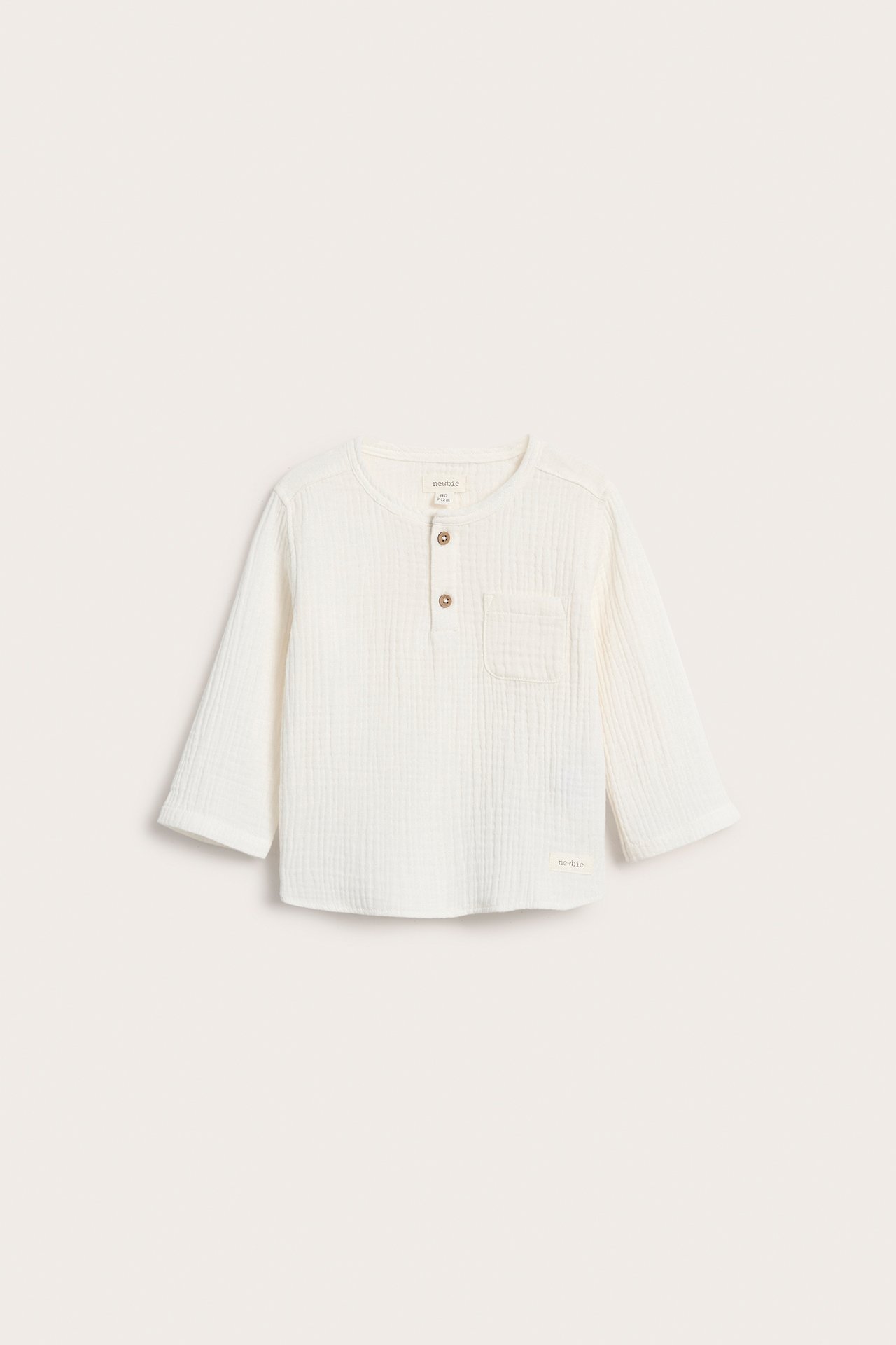 Vorderansicht eines cremefarbenen Kinder-Langarmshirts aus Baumwollmusselin mit zwei Knöpfen und Brusttasche.