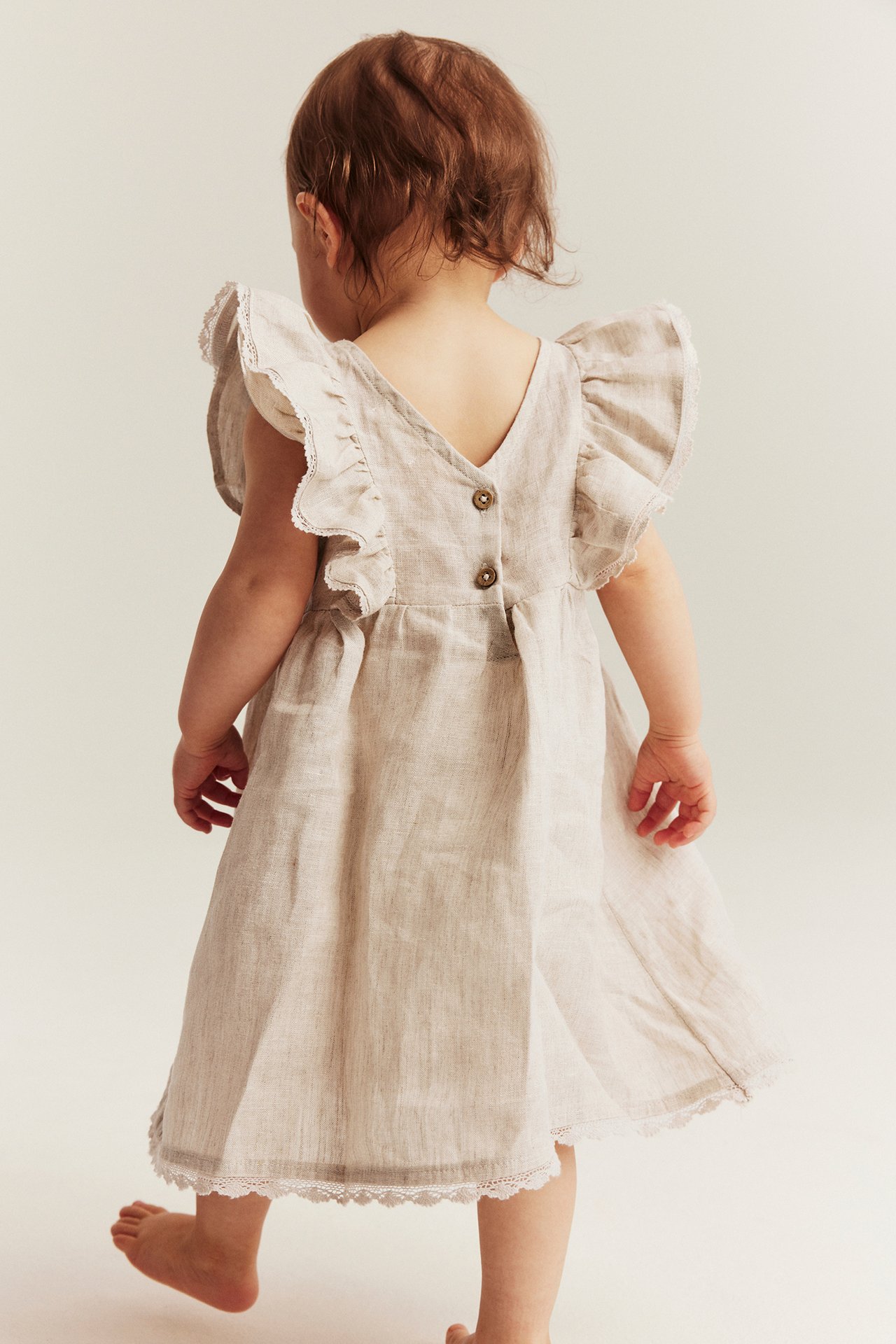 Rückenansicht eines Babys in hellbeigem Leinenkleid mit Rüschenärmeln, Spitzenbesatz und Knöpfen hinten.