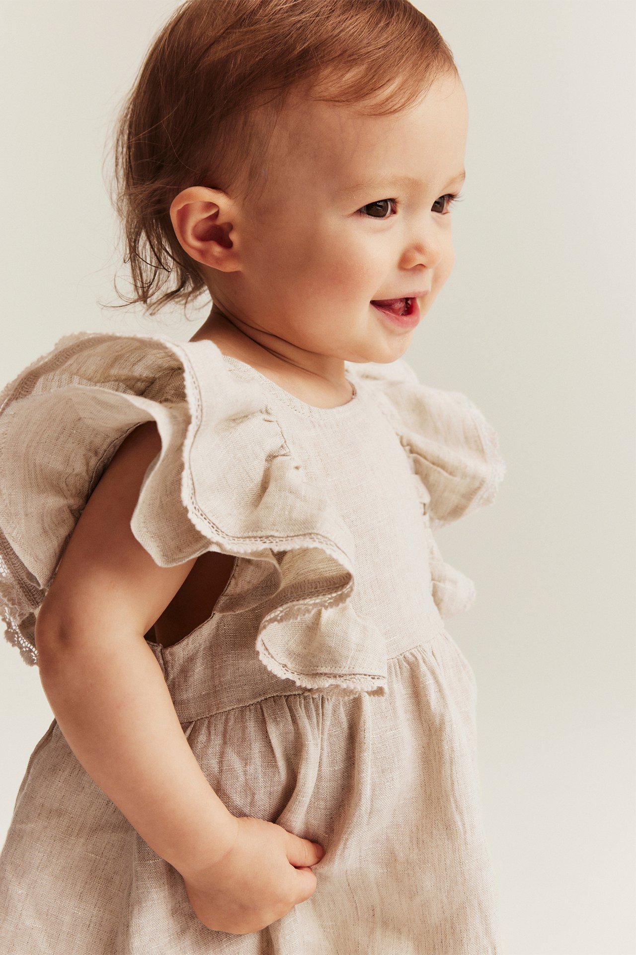 Side view of a smiling baby wearing a light beige linen dress with ruffled sleeves and lace trim.
