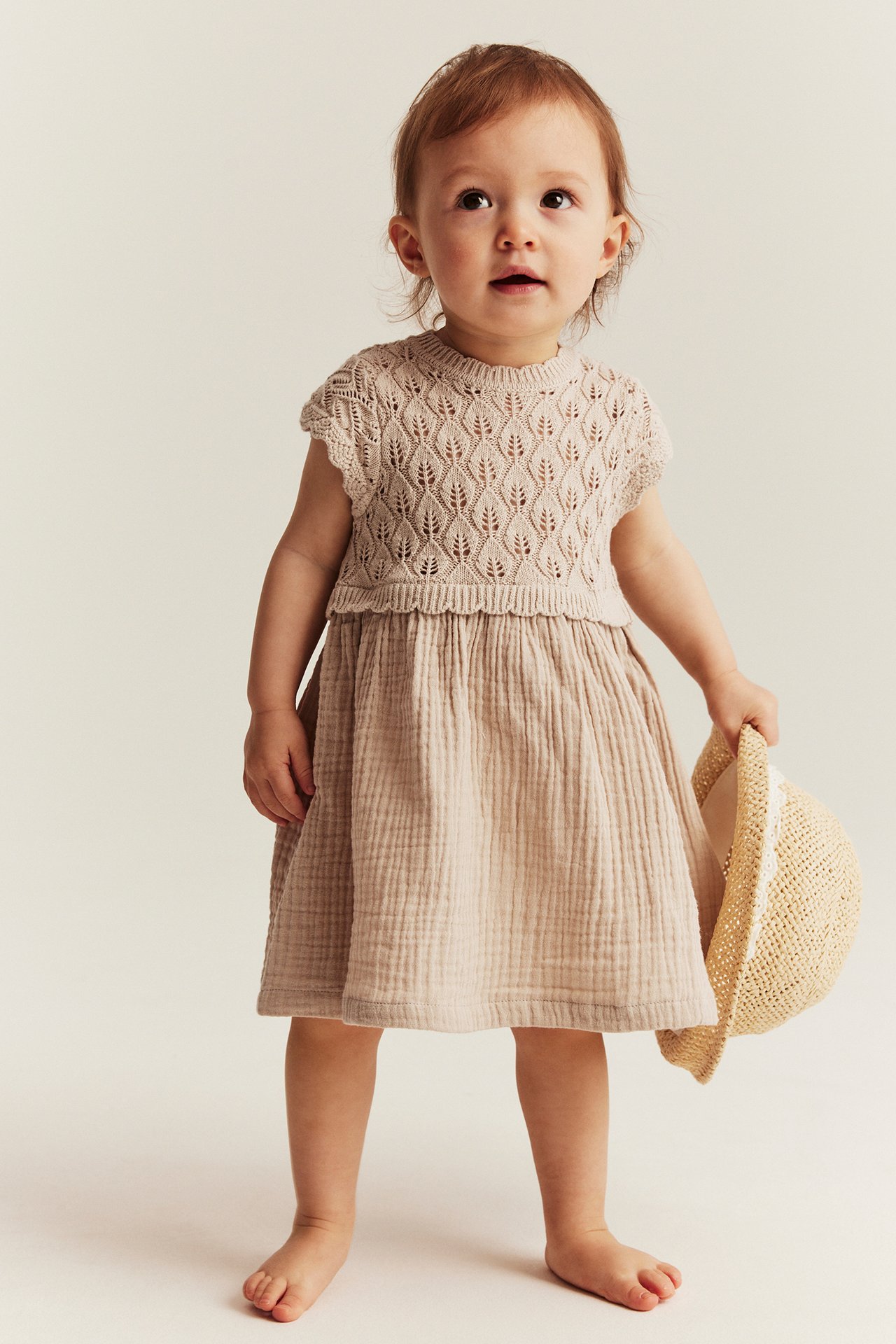 Vorderansicht: Baby in beigem Strickkleid mit Blattmuster-Oberteil und Musselinrock, hält Strohhut.