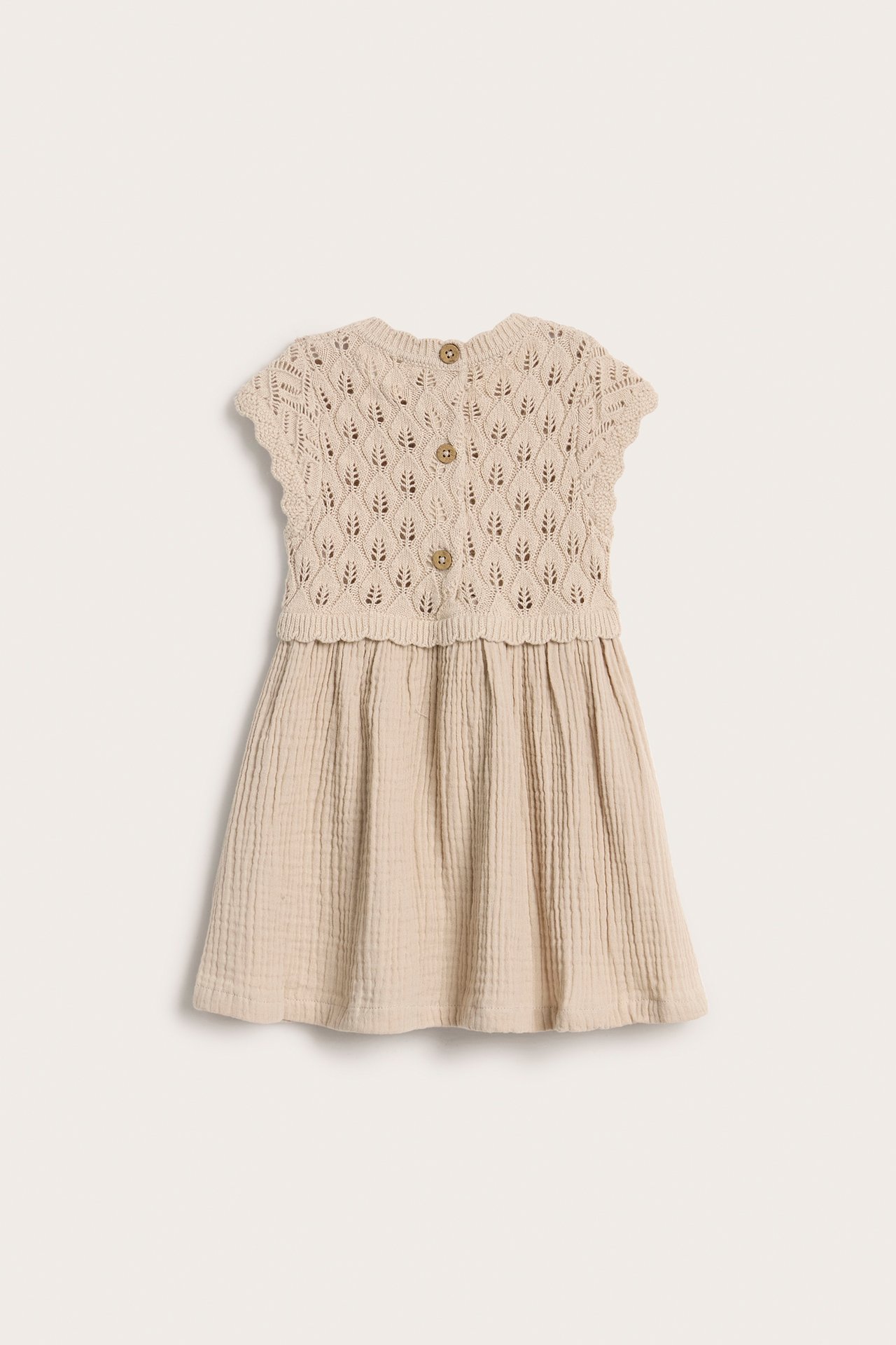 Vorderansicht: beiges Babykleid mit gestricktem, gemustertem Oberteil, schlichtem Musselinrock und Knöpfen vorne.