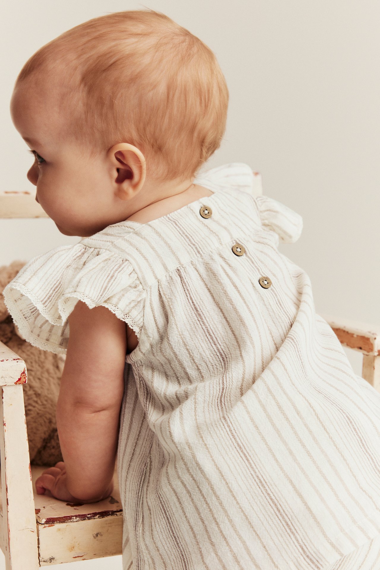 Rückansicht, Baby in gestreiftem Off-White Baumwolltop mit Rüschenärmeln, Spitze und Knöpfen am Rücken.