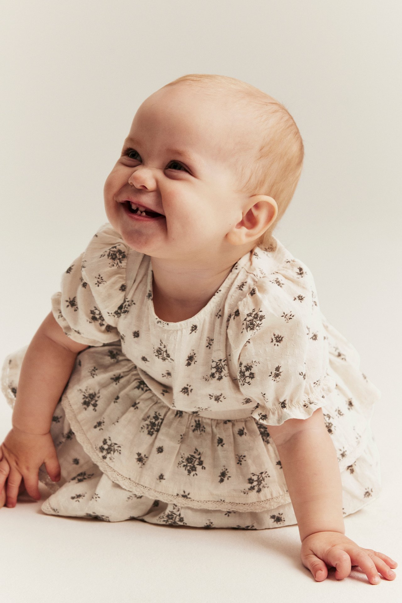 Front-side view of a smiling baby wearing a light beige floral cotton playsuit with ruffles and elasticated sleeves.