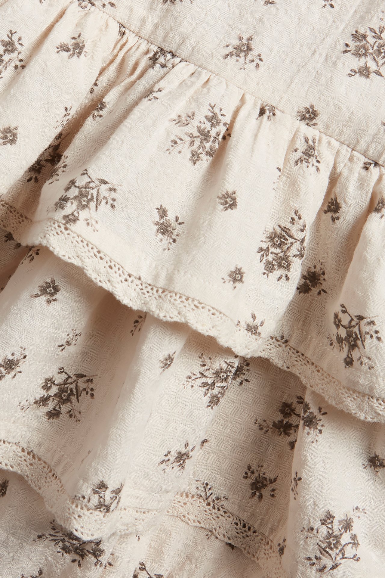 Close-up of a light beige cotton fabric with small brown floral print, ruffles, and lace trim.