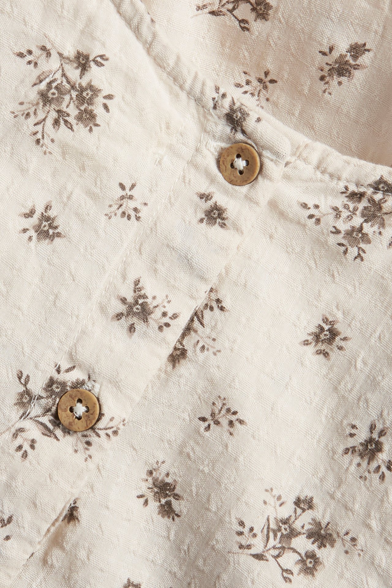 Close-up of a light beige woven cotton playsuit with a small brown floral pattern and two wooden buttons.