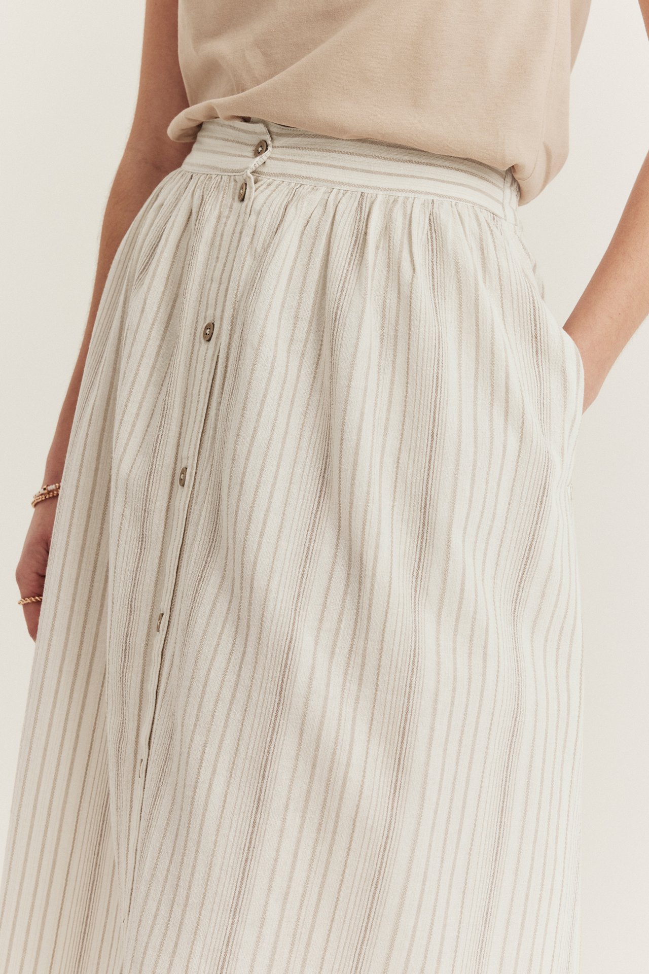 Front view: woman in offwhite striped cotton skirt with button front, hand in pocket, and beige t-shirt.