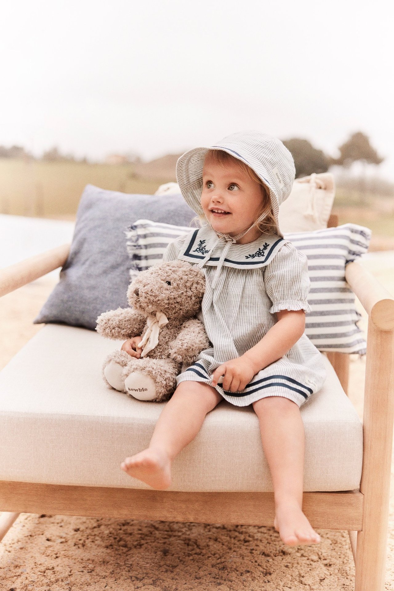 Vorderansicht: Kind in gestreiftem Baumwollkleid mit Matrosenkragen, passendem Hut, hält braunen Teddybär.