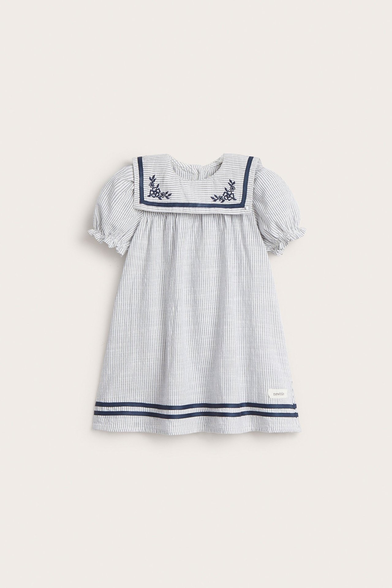 Vorderansicht eines gestreiften Baumwollkleides für Kinder in Off-White mit marineblauem Matrosenkragen, Stickerei, Puffärmeln und Saumbändern.