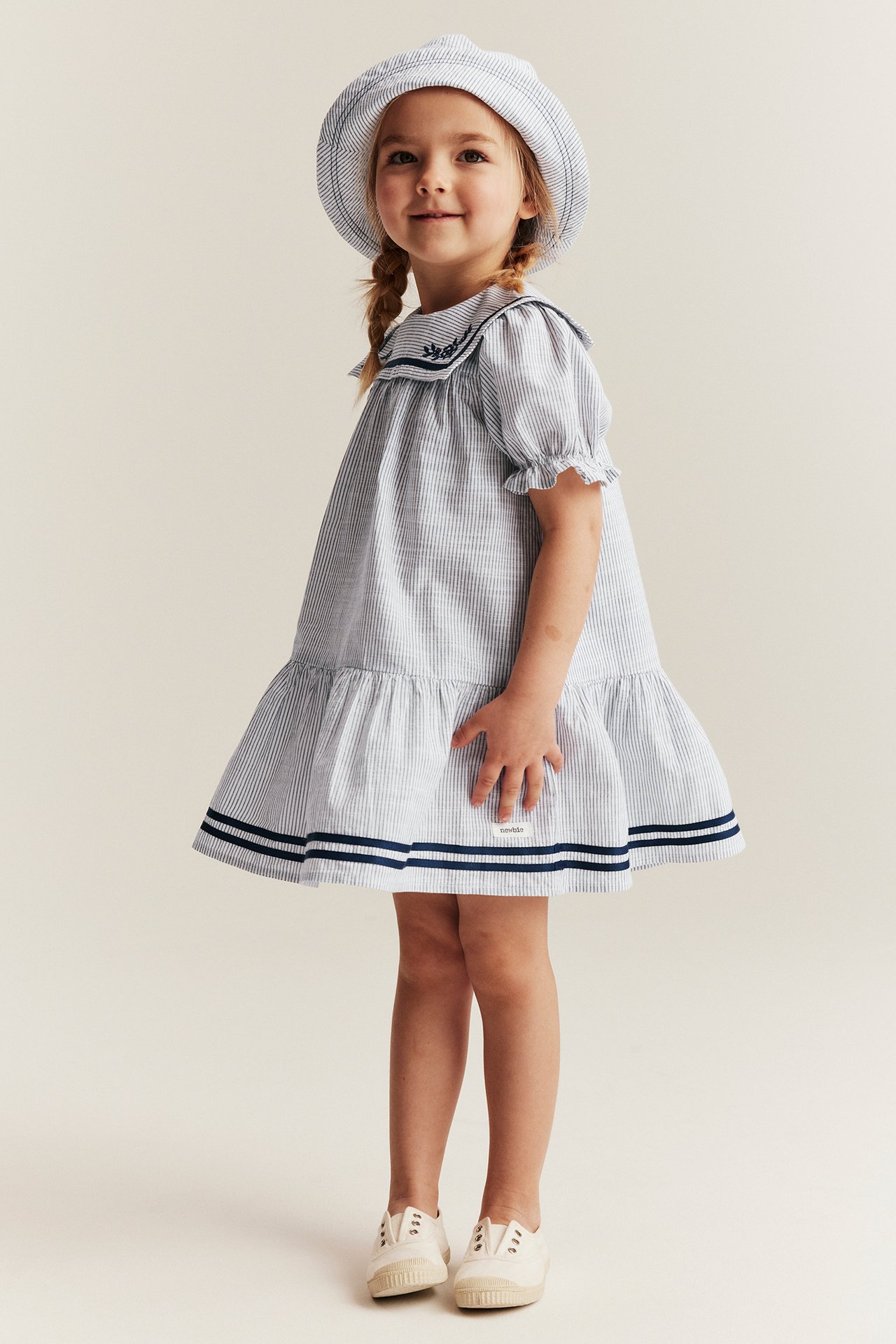 Vorderansicht: Kind in einem cremefarbenen gestreiften Kleid mit blauem Matrosenkragen, passendem Hut und weißen Schuhen.