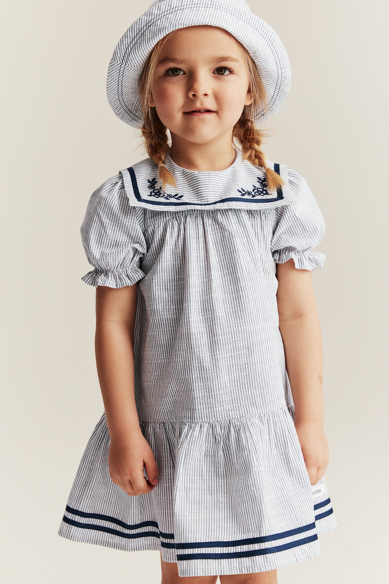 Vorderansicht: Kind in off-white gestreiftem Baumwollkleid mit besticktem Matrosenkragen, Puffärmeln und passendem Hut.
