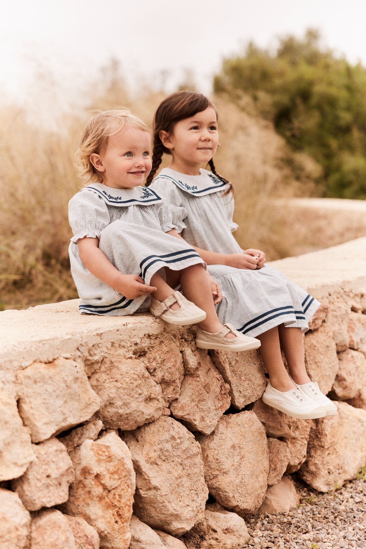 Zwei Kinder tragen gestreifte Baumwollkleider in Off-White mit marineblauen Matrosenkragen. Eines hat beige, eines weiße Schuhe.