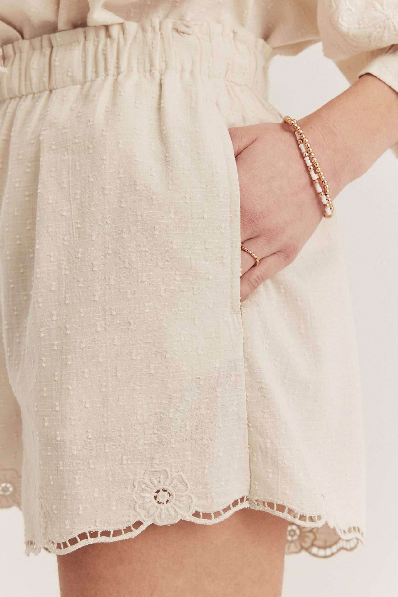 Close-up of woman wearing light beige woven shorts with dotted texture, elastic waist, embroidered hem, and beaded bracelets.