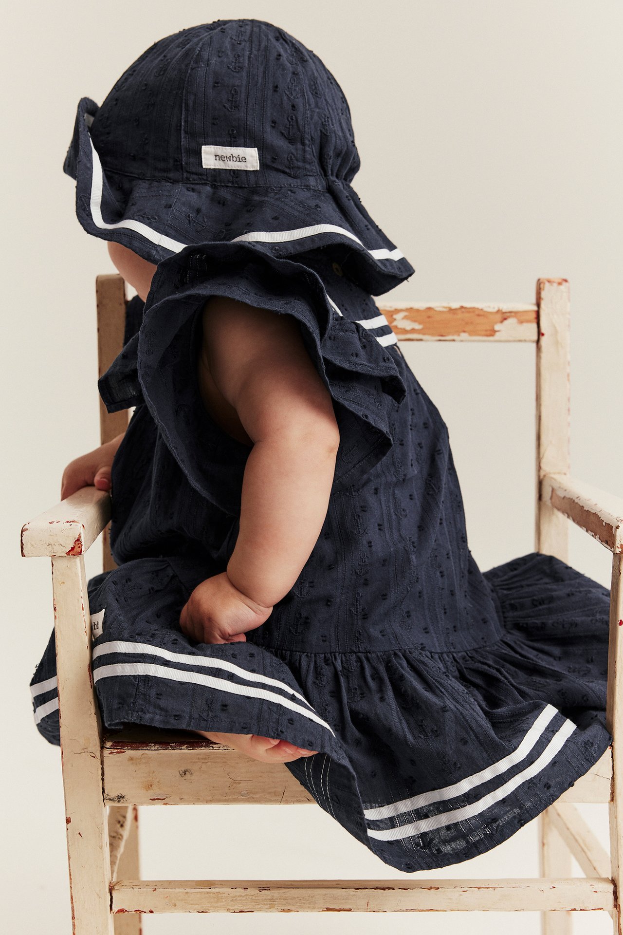 Rückenansicht eines Babys in dunkelblauem Bio-Baumwollkleid mit Rüschenärmeln und weißen Streifen, passendem Hut.