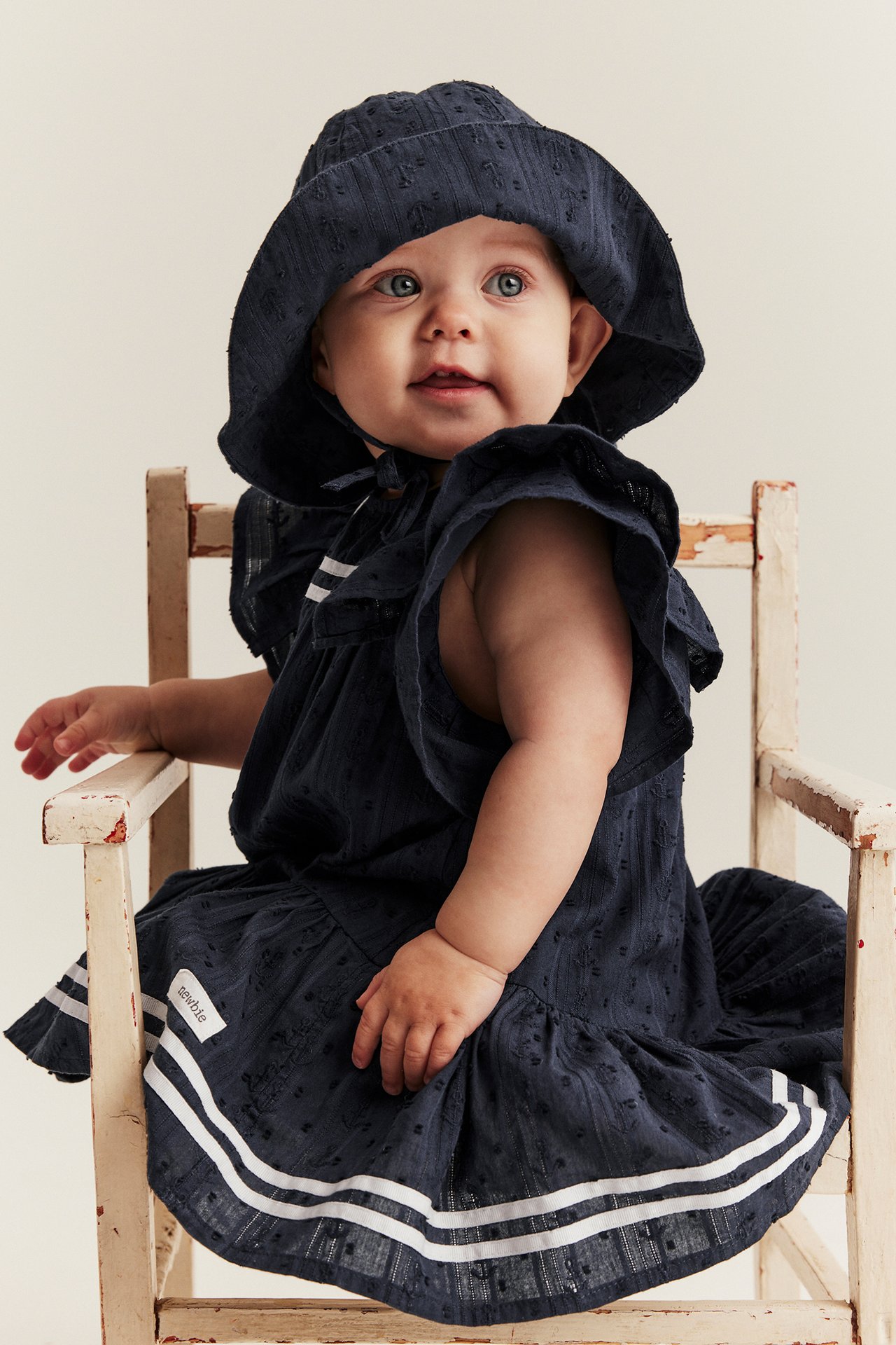 Vorderansicht: Baby in dunkelblauem Dobby-Baumwollkleid mit Rüschenärmeln, weißen Streifen und passendem Hut.
