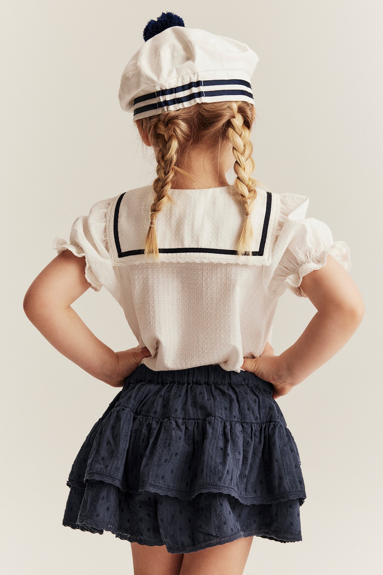 Back view of a kid in a dark blue ruffled skort, white sailor blouse, and white sailor hat.