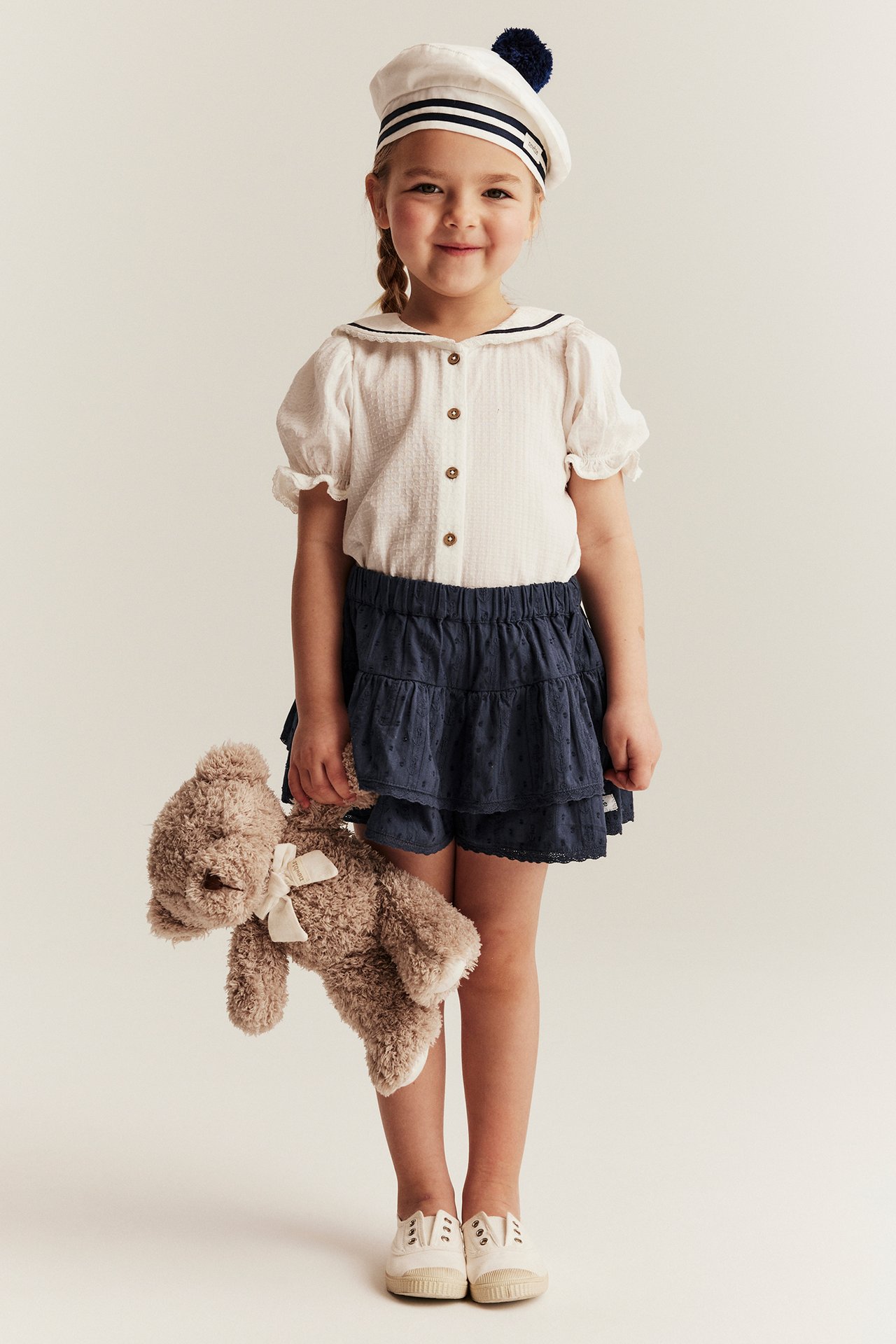 Kid in a dark blue ruffled skort, white blouse, sailor hat, and white shoes, holding a teddy bear.