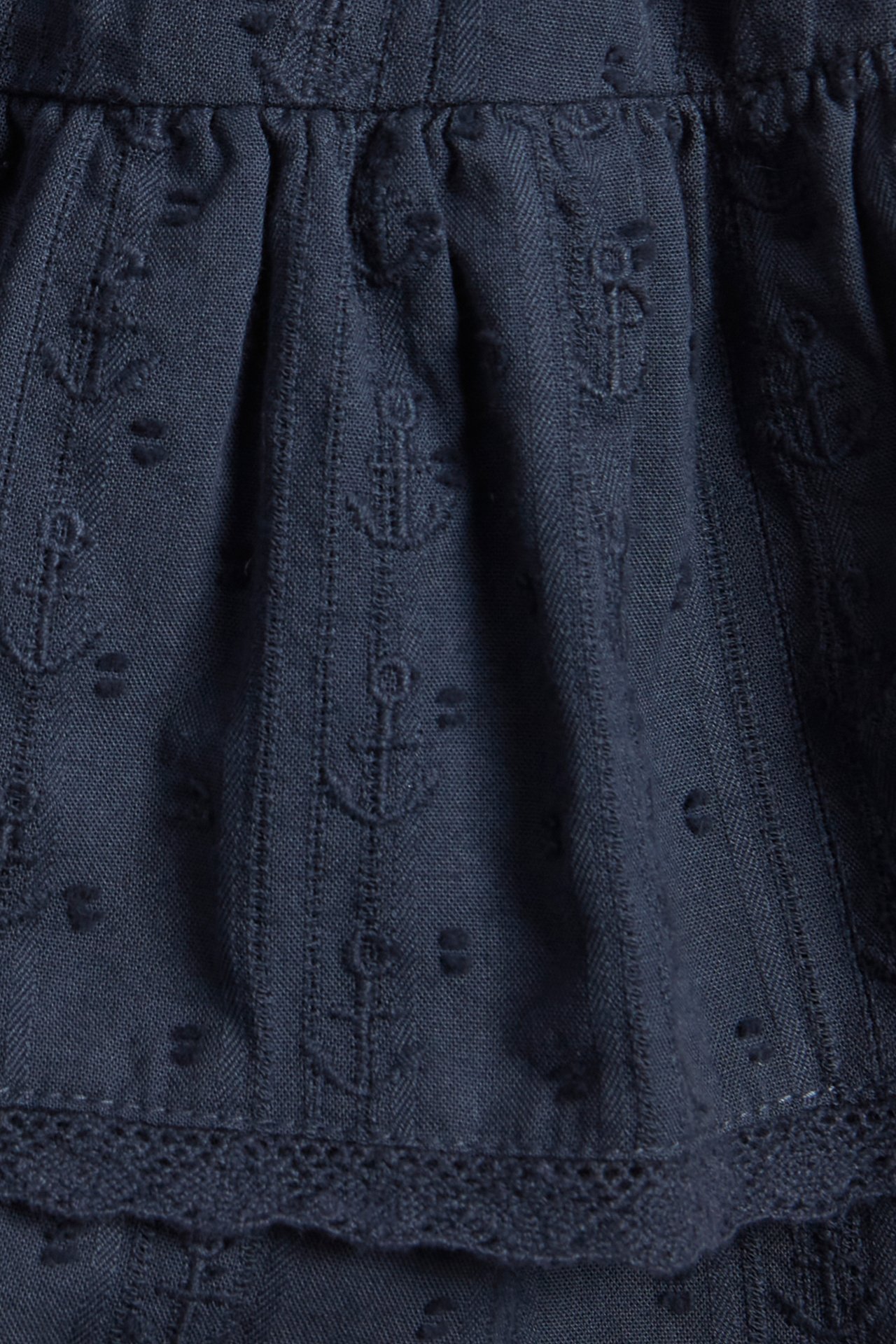 Close-up of a kid's dark blue cotton skort with embroidered anchors, dobby stripes, ruffles, and a lace hem.