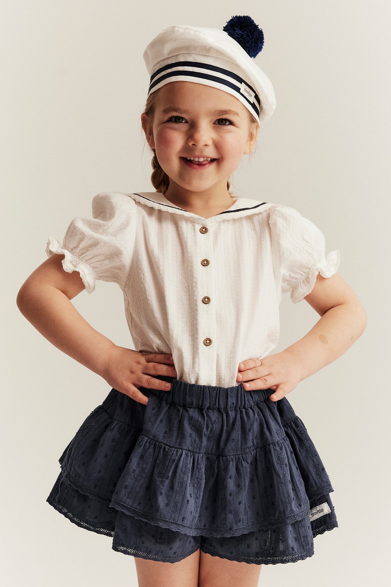 Front view: Kid in dark blue cotton ruffled shorts-skirt, white sailor blouse, and white sailor hat.