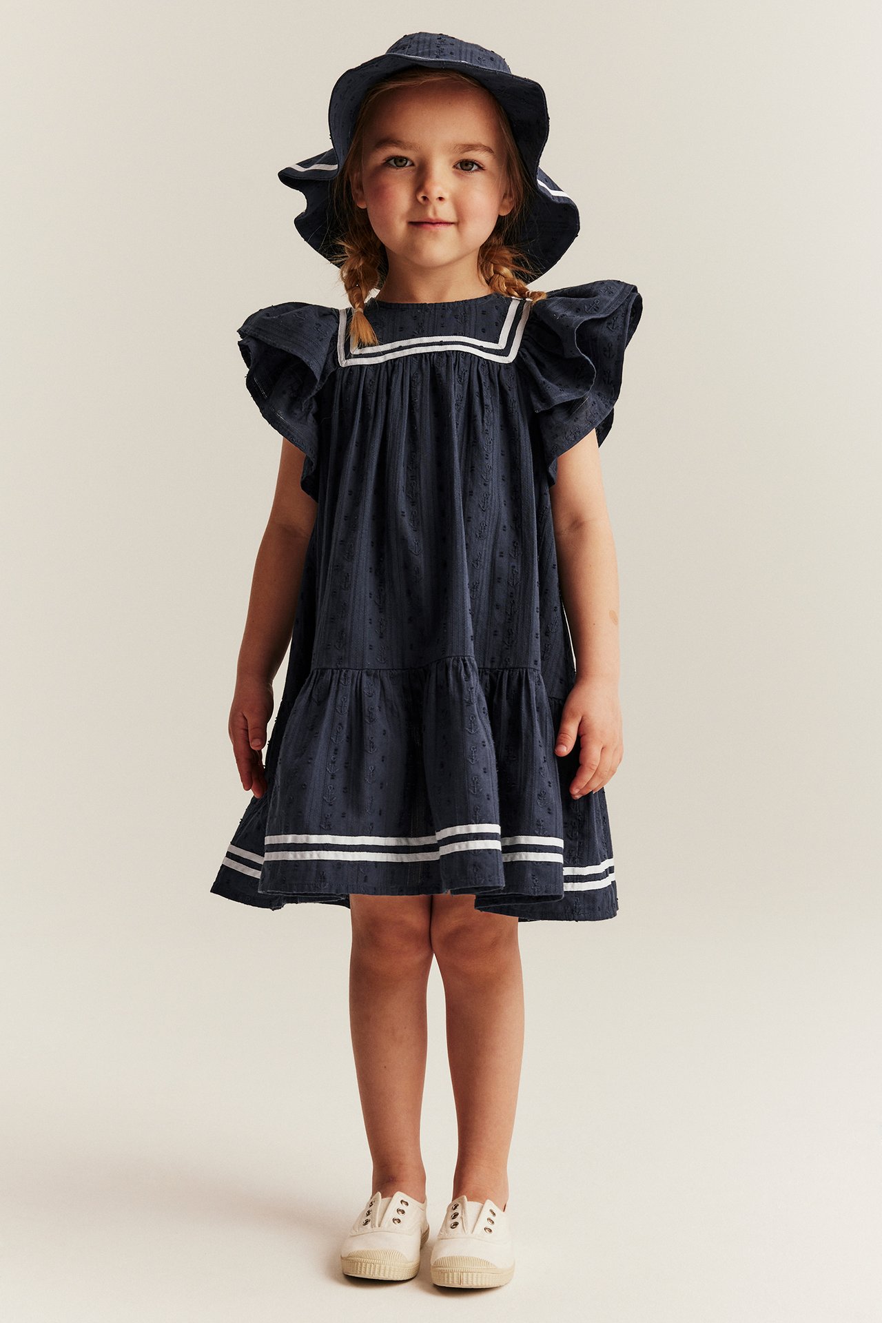 Kind in dunkelblauem Dobby-Baumwollkleid mit Rüschenärmeln, weißen Streifen, passendem Hut und cremefarbenen Schuhen.
