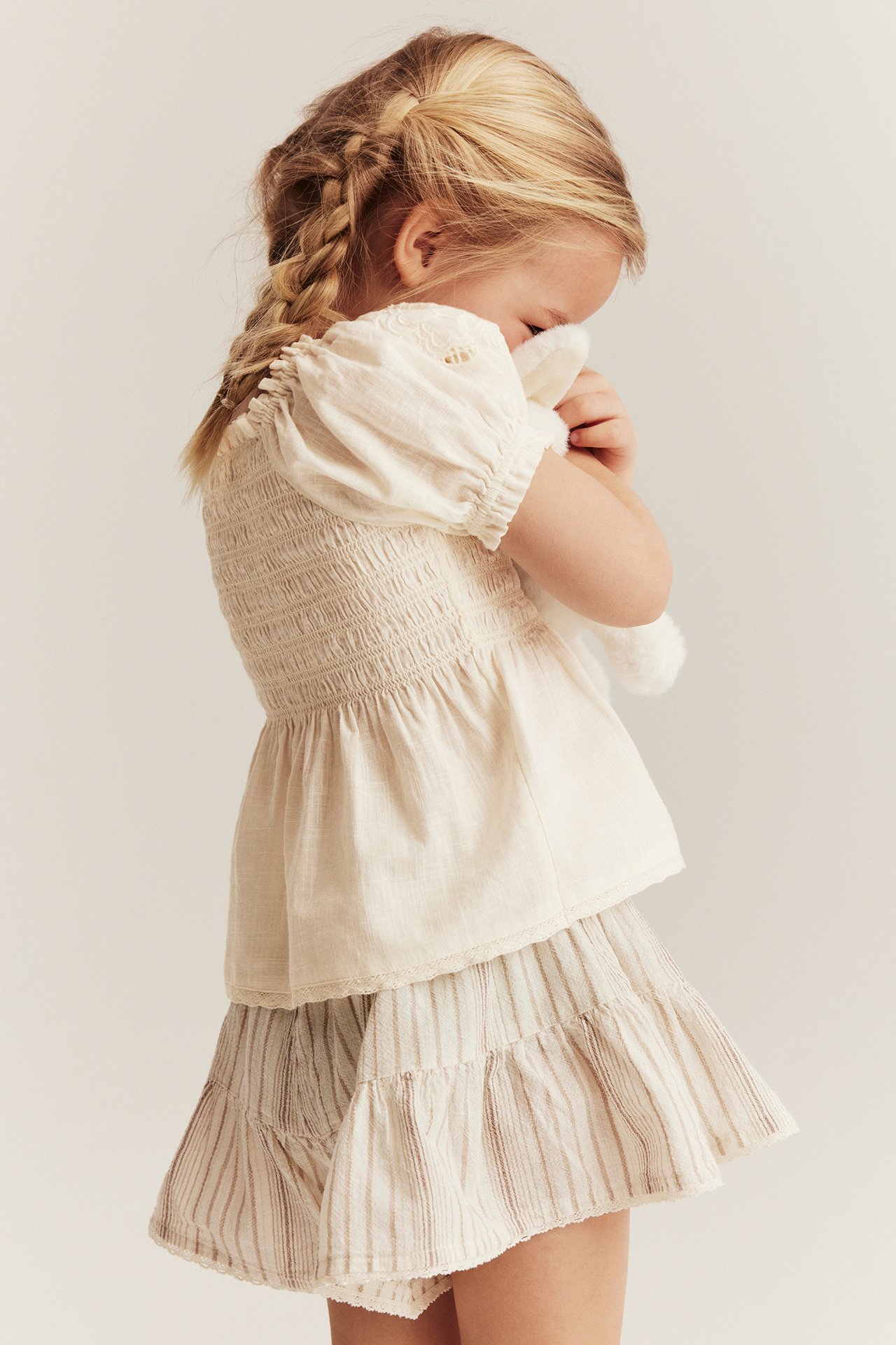 Kid in off-white cotton blouse with smock, puff sleeves, and striped skirt, holding a white toy.