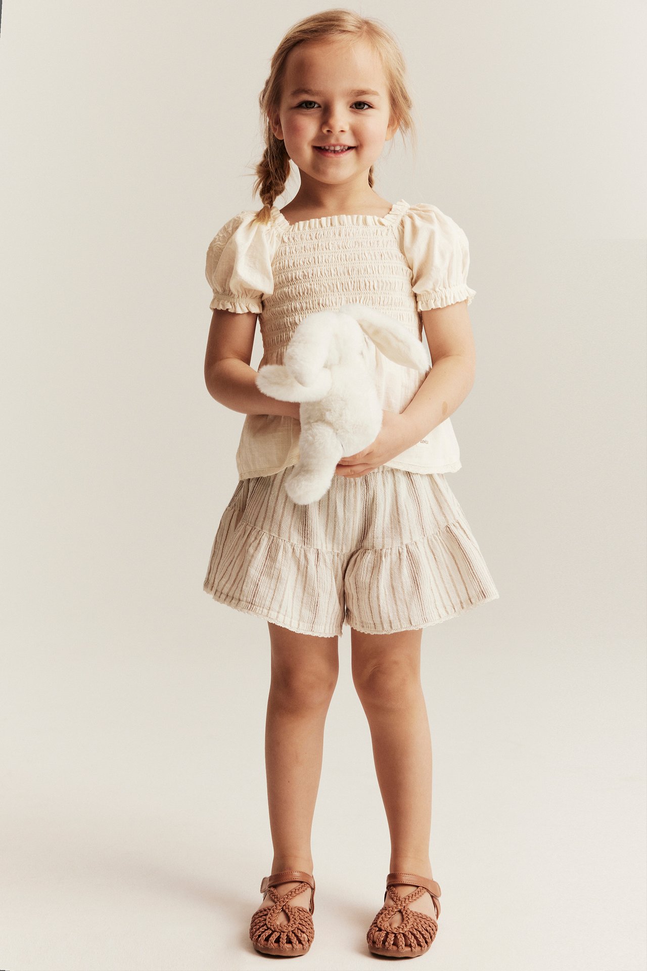 Front view: Kid in off-white smocked cotton blouse, striped shorts, brown sandals, holding white bunny.