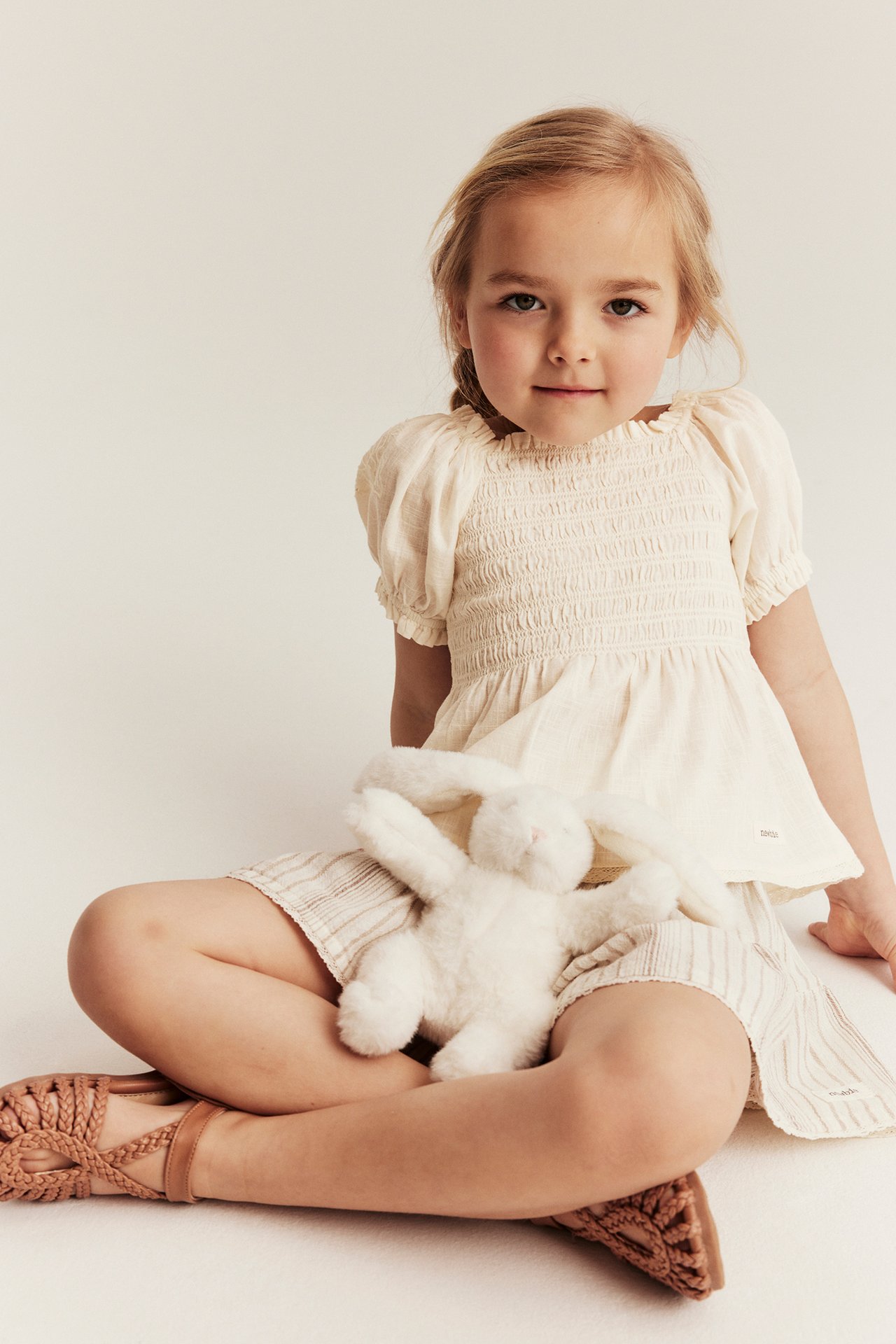 Front view of a kid in an off-white smocked cotton blouse, striped shorts, braided sandals, holding a white bunny.