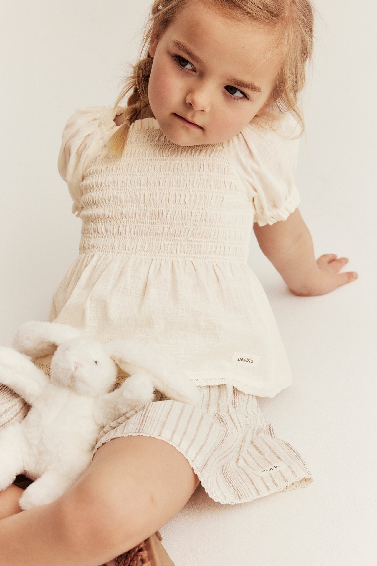 Front view of a kid in an offwhite smocked cotton blouse, striped shorts, holding a white bunny.