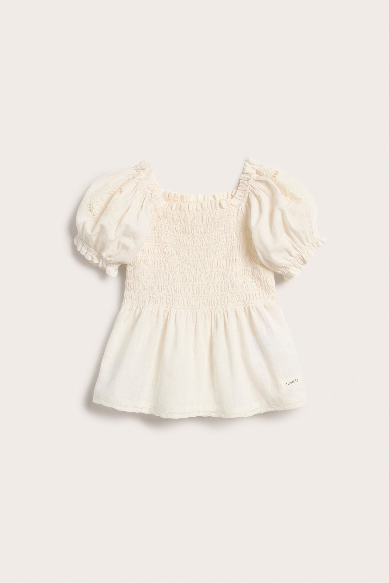 Front view of a kid's off-white cotton blouse with smocking, embroidered puff sleeves, and a lace-trimmed peplum.