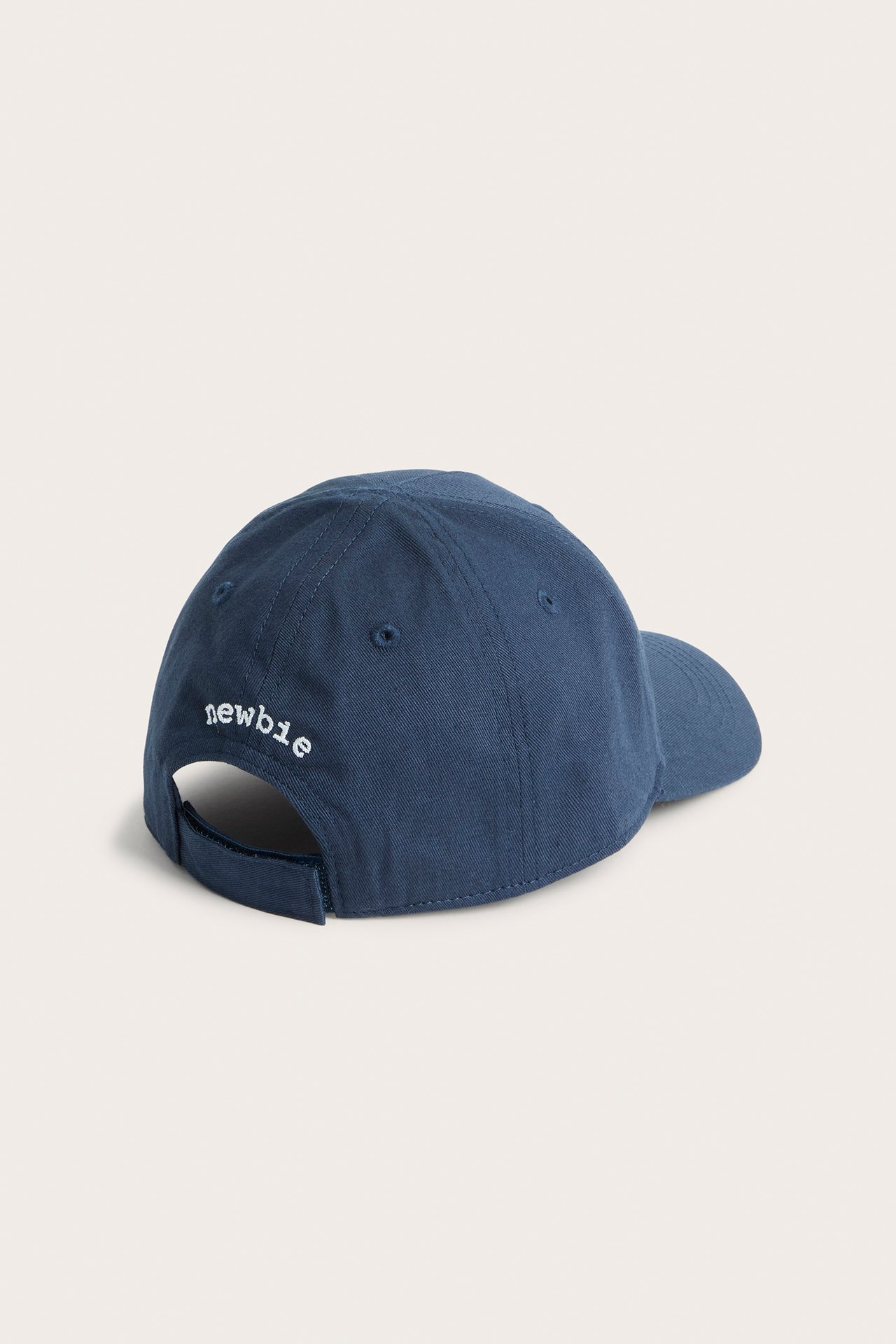 Back-side view of a kid's dark blue cotton cap with white "newbie" embroidery and an adjustable strap.