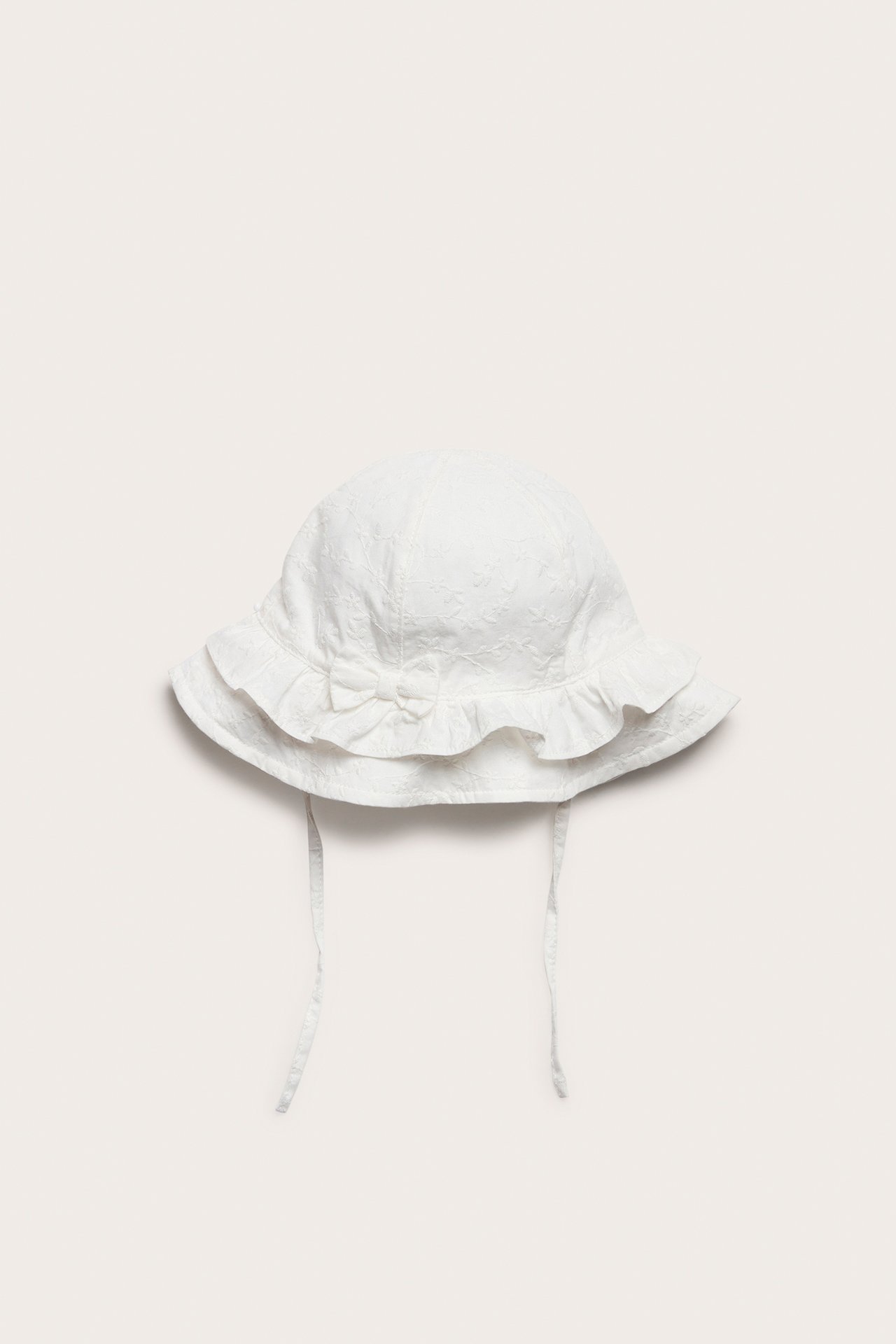 Front view: Offwhite baby sun hat in embroidered fabric with ruffled brim, small bow, and tie-straps.