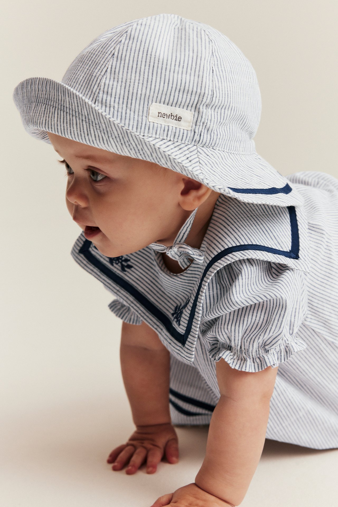 Side view of a baby wearing an off-white striped sun hat with blue ribbon and a matching striped dress.