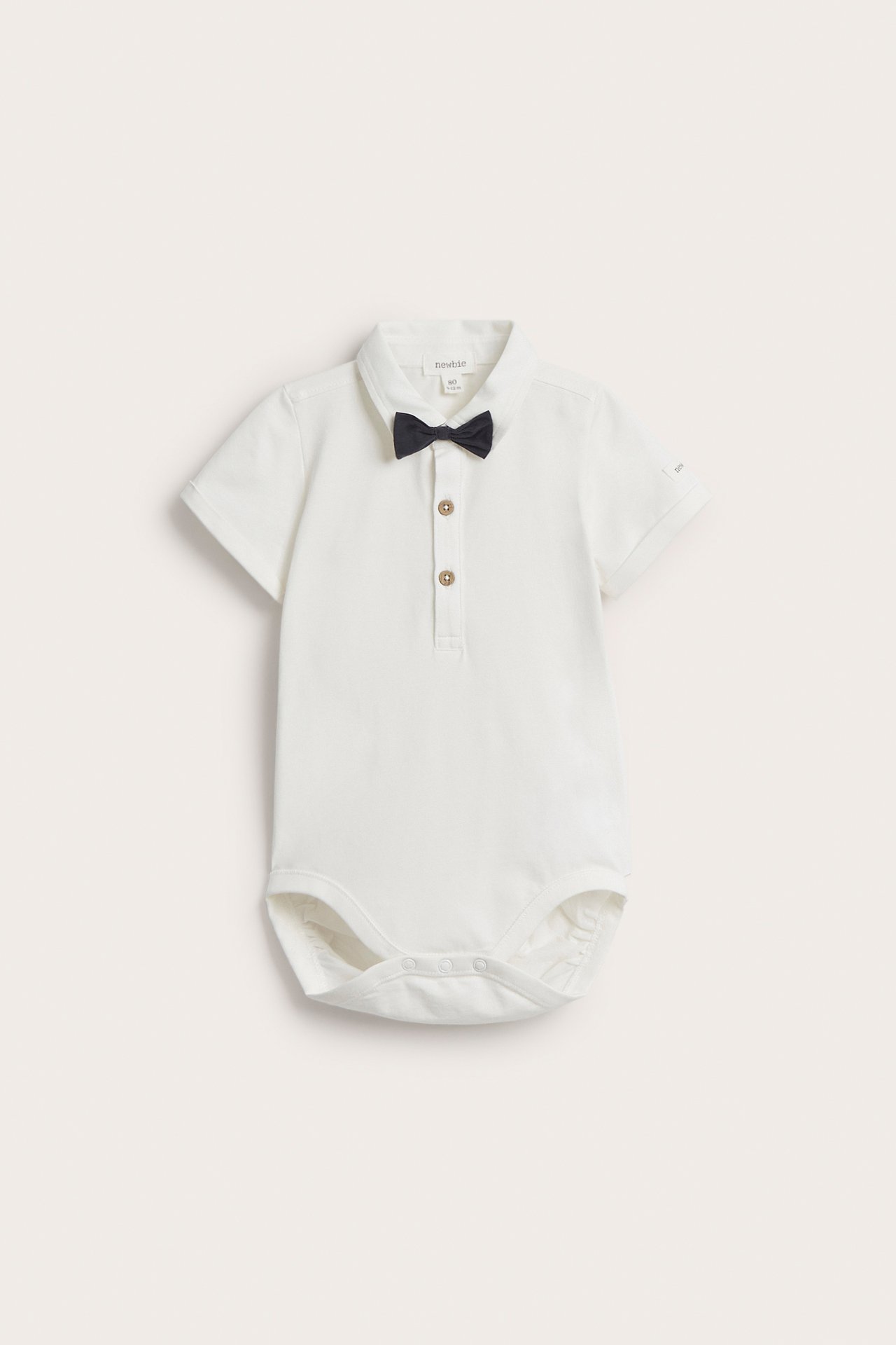 Vorderansicht: Off-white kurzärmeliger Baby-Body mit Kragen, Knöpfen vorne, Druckknöpfen im Schritt und dunkler Fliege.