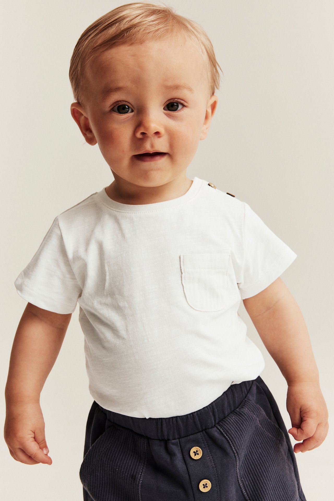 Front view of a baby wearing an off-white cotton short-sleeved bodysuit with a chest pocket and dark blue shorts.