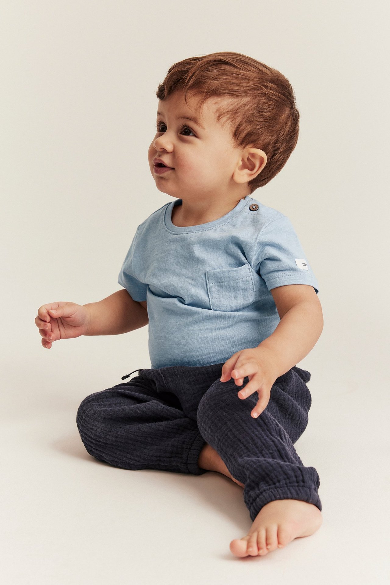 Front-side view of a baby in a light blue slub jersey bodysuit with pocket and dark blue pants.