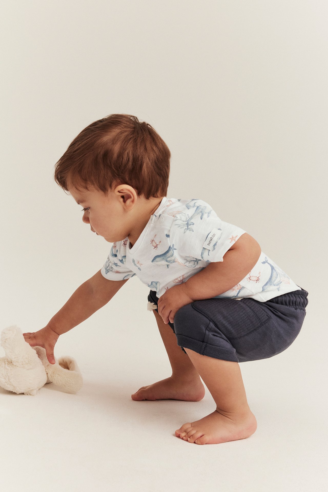 Side view of a baby in an off-white animal-patterned cotton bodysuit and dark blue shorts, reaching for a toy.