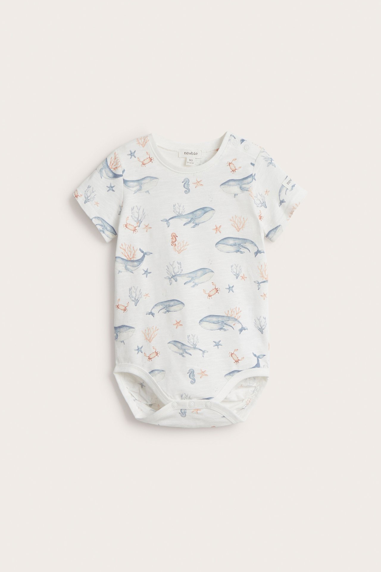 Front view: off-white baby short-sleeved cotton bodysuit with whale and coral pattern, snap buttons.