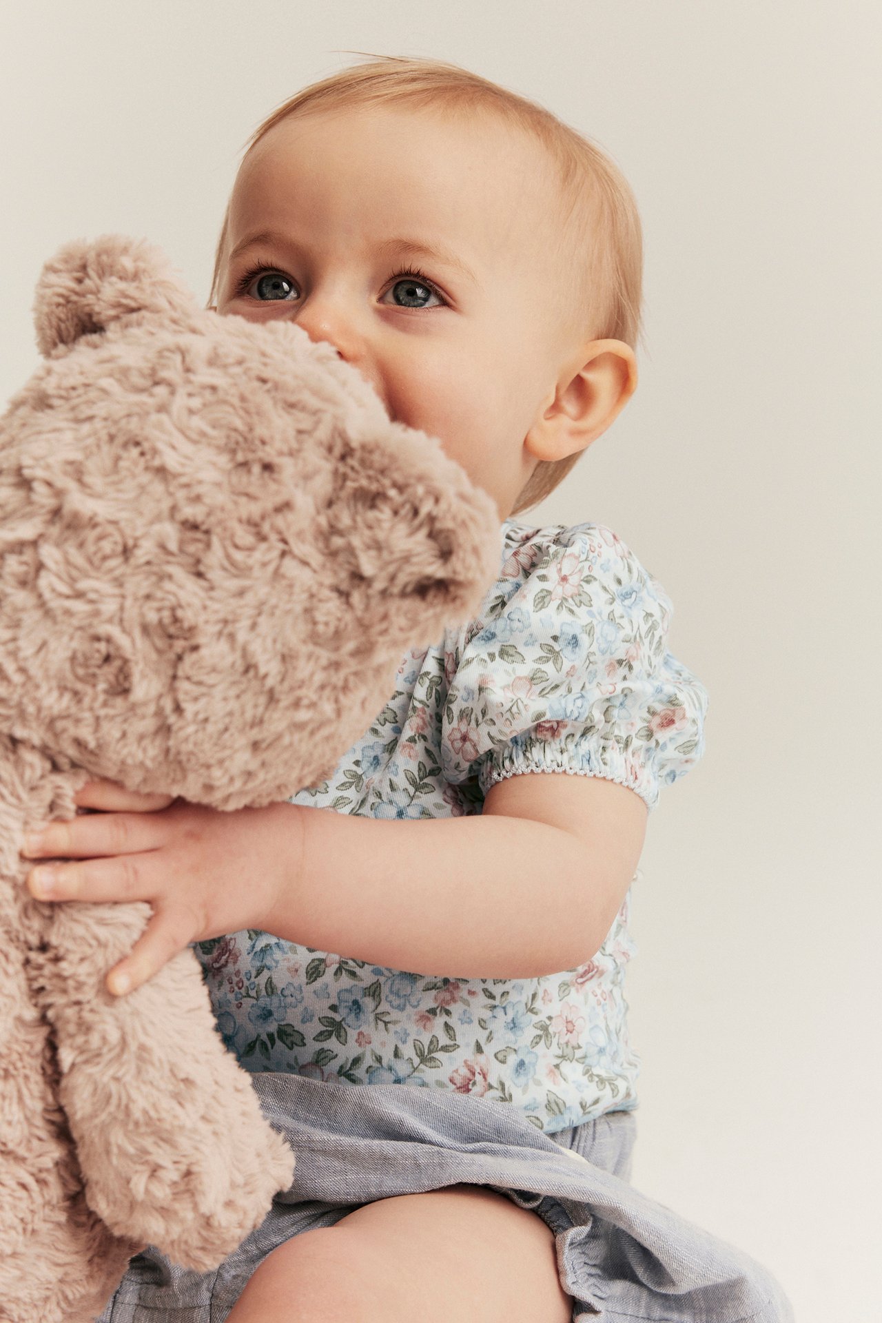Nahaufnahme eines Babys in einem cremefarbenen, geblümten Kurzarm-Body, hellblauen Shorts, hält einen braunen Teddybär.