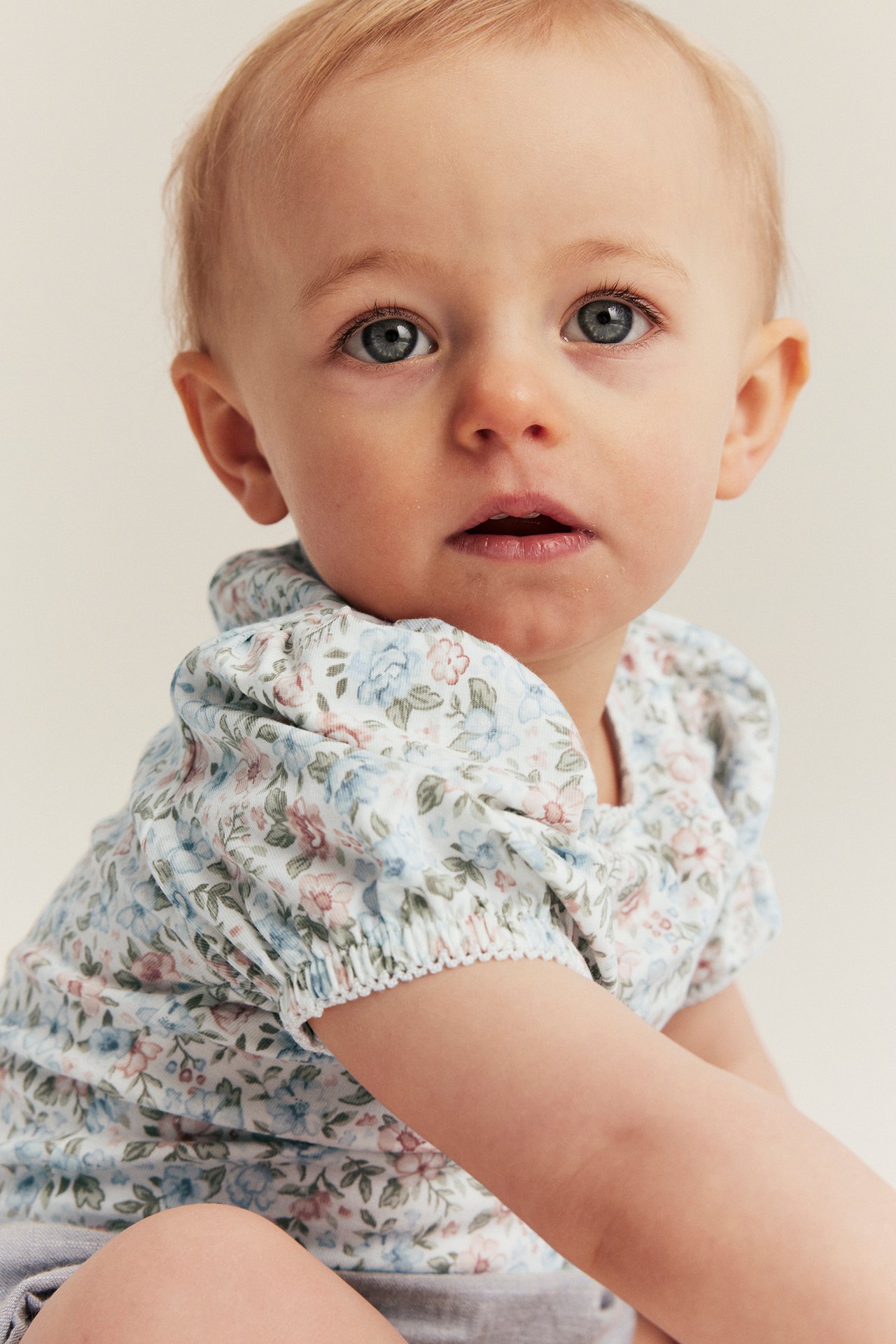 Nahaufnahme eines Babys in einem cremeweißen, geblümten Kurzarm-Body mit Puffärmeln und Gummizug.