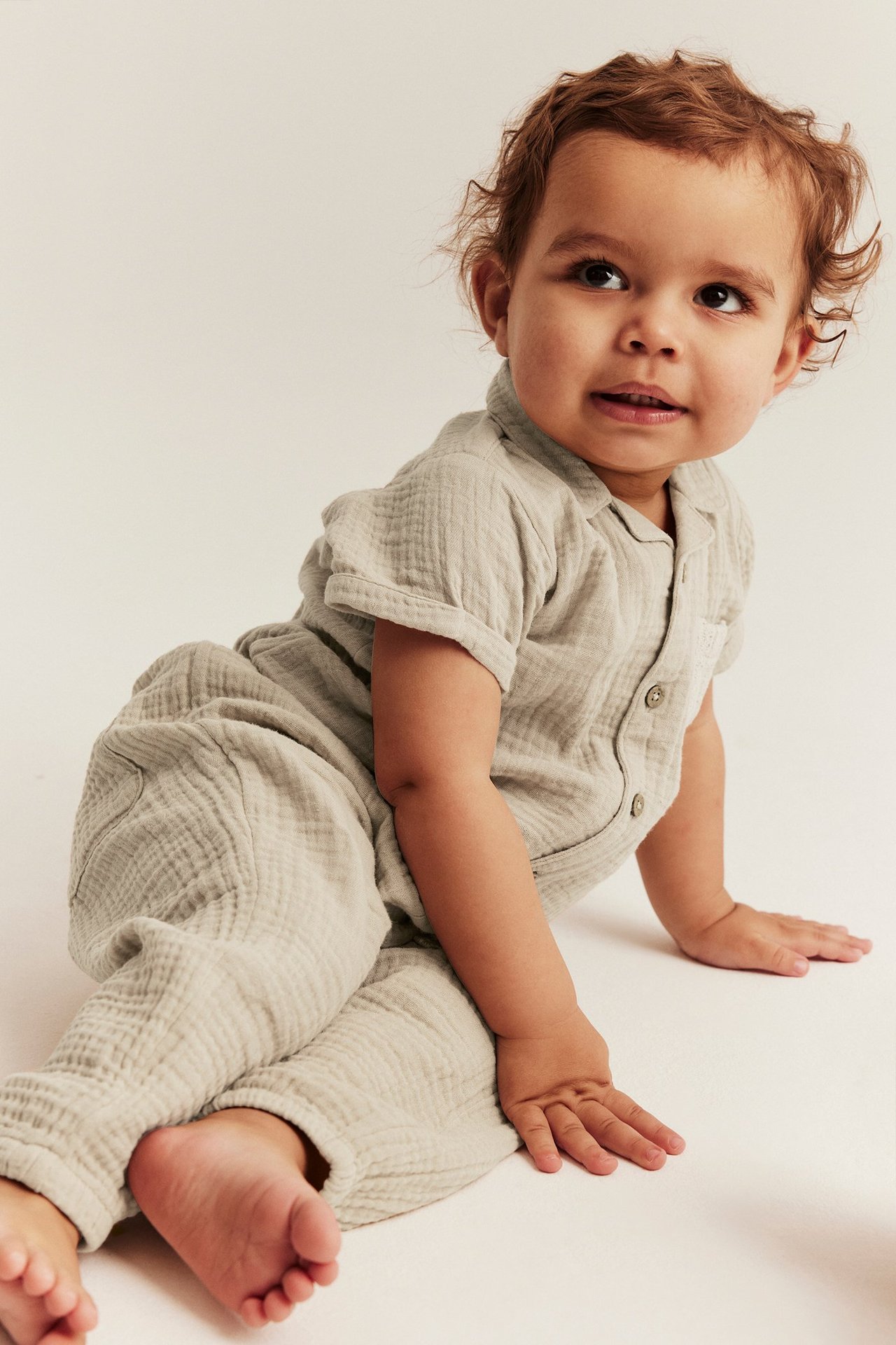 Front-side view: baby in a beige cotton muslin short-sleeved body with buttons and matching pants.