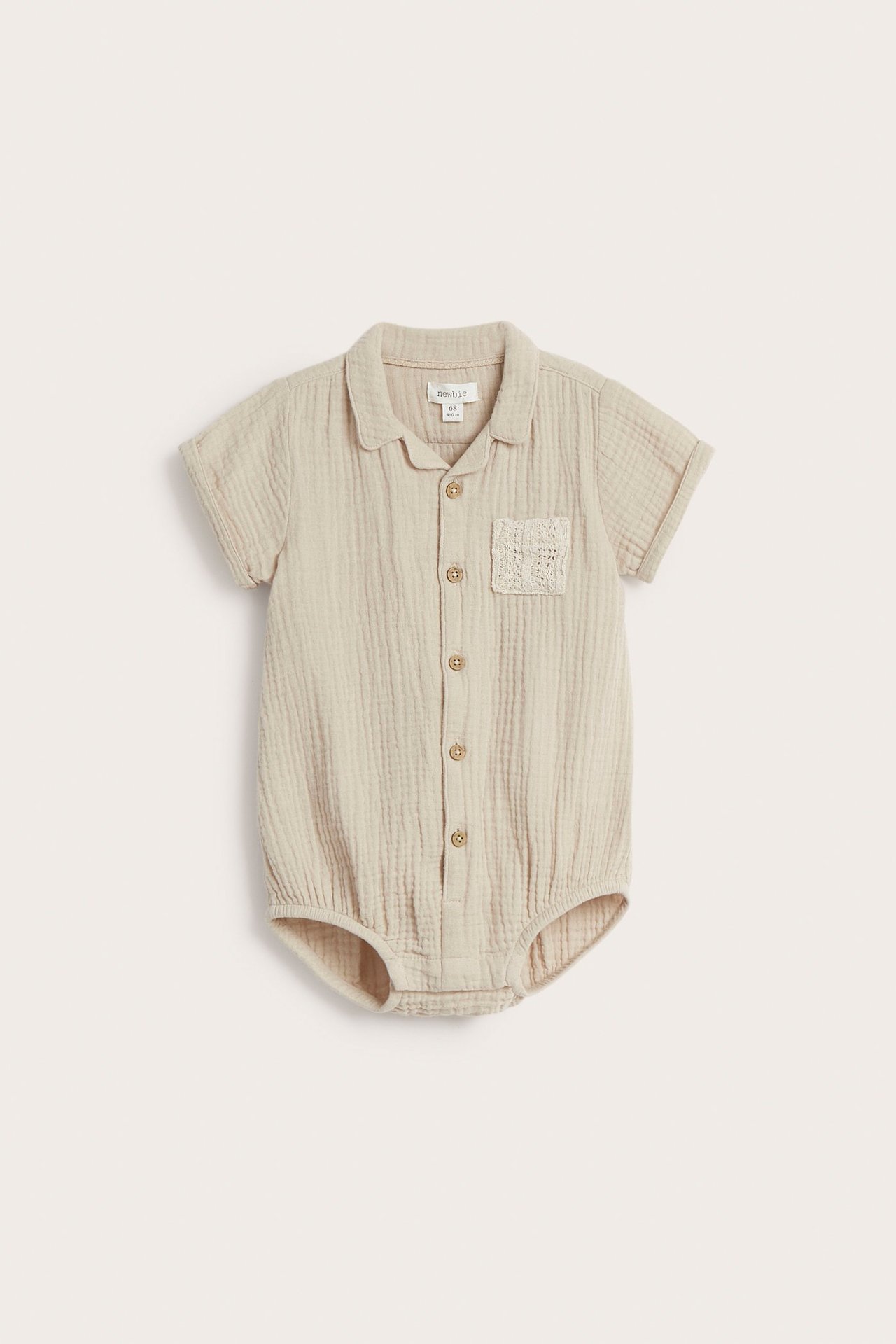 Front view of a beige cotton muslin baby bodysuit with front buttons and a textured chest pocket.